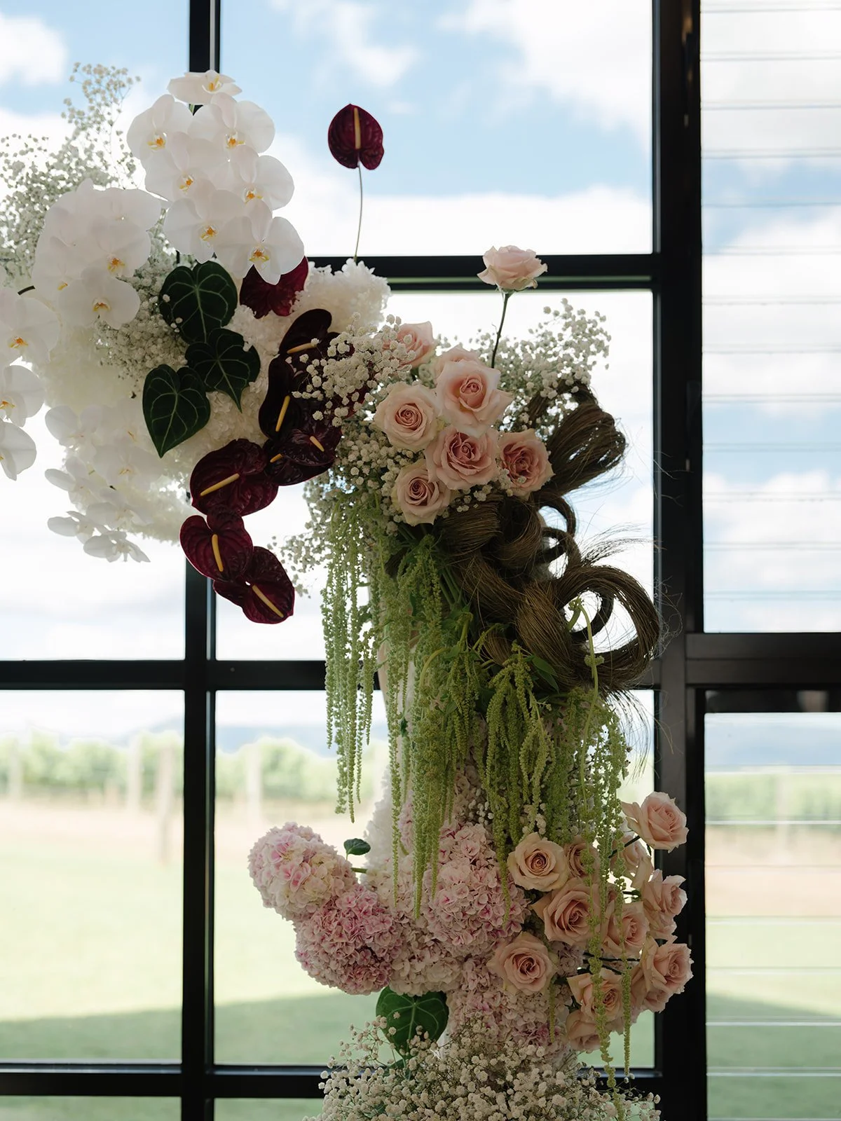 A floral arrangement featuring white orchids, pink roses, burgundy anthuriums, and various green foliage, set against large windows showing a cloudy sky and distant landscape.