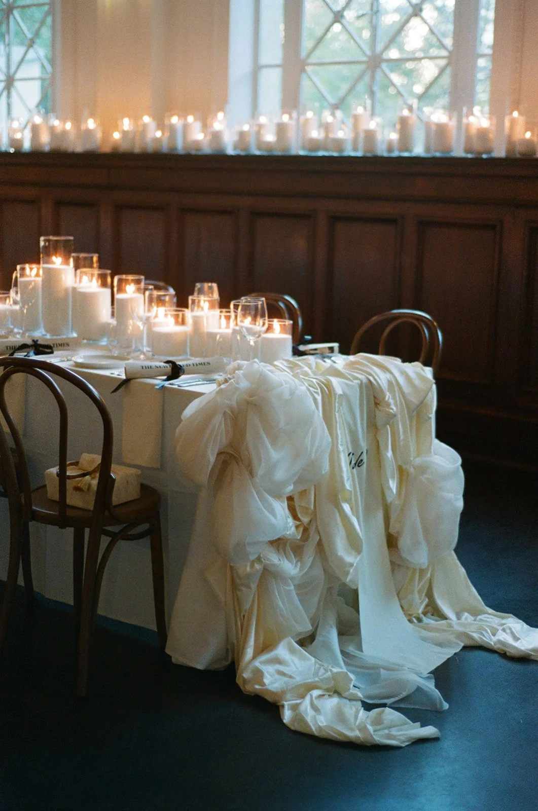 A wedding table setting with white candles, wine glasses, and a cream-colored fabric draped over the side, in a room with wooden paneling and large windows.