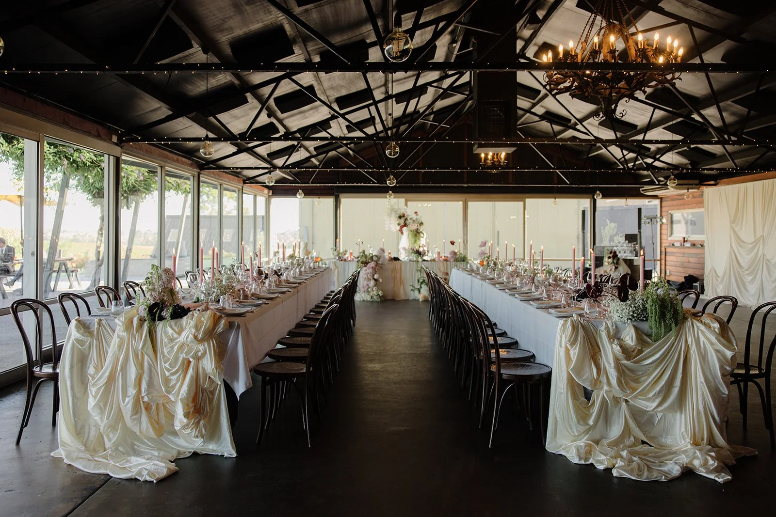 Elegant indoor wedding reception with long tables decorated with candles, flowers, and drapery, with large windows and chandeliers overhead.