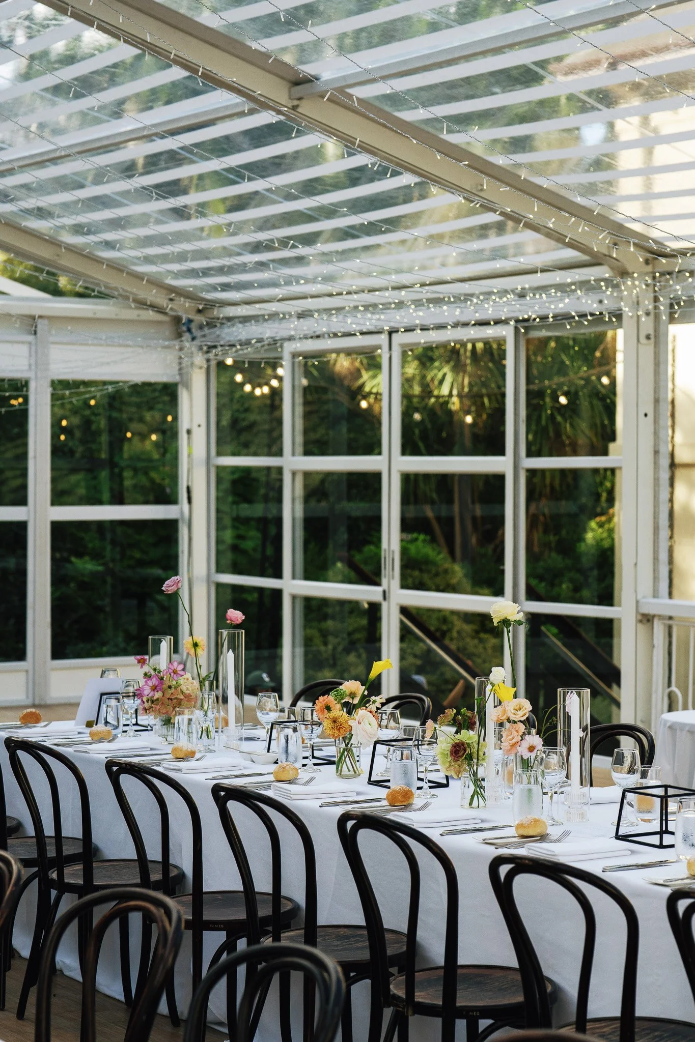 A long banquet table decorated with floral centerpieces, glassware, and bread rolls, set for a formal event inside a glass-enclosed venue with string lights and greenery outside.