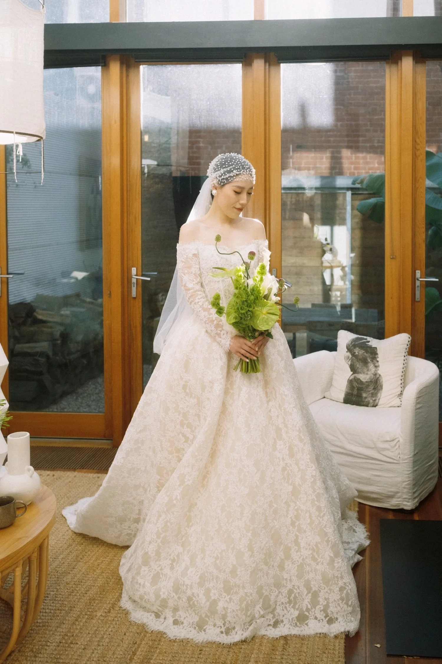 A bride in a wedding dress holding a bouquet of flowers, standing indoors in front of glass doors with wooden frames, in a cozy room with a white armchair and decorative pillows.