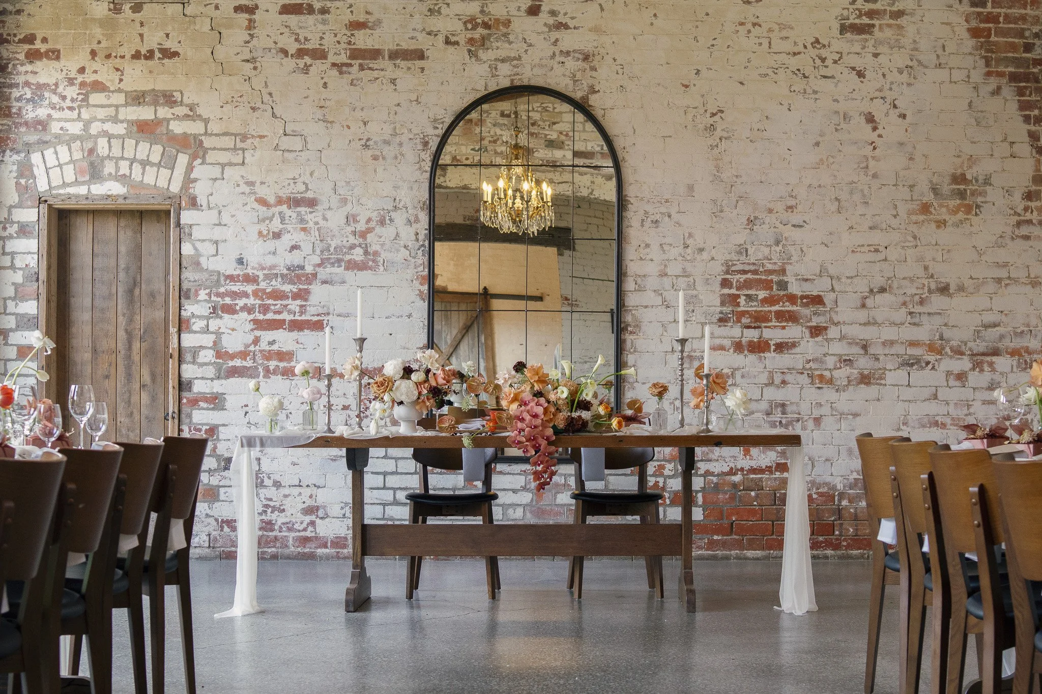 An indoor event space with a rustic exposed brick wall, a large arched mirror, a chandelier reflected in the mirror, and a long table decorated with a floral centerpiece and candles.
