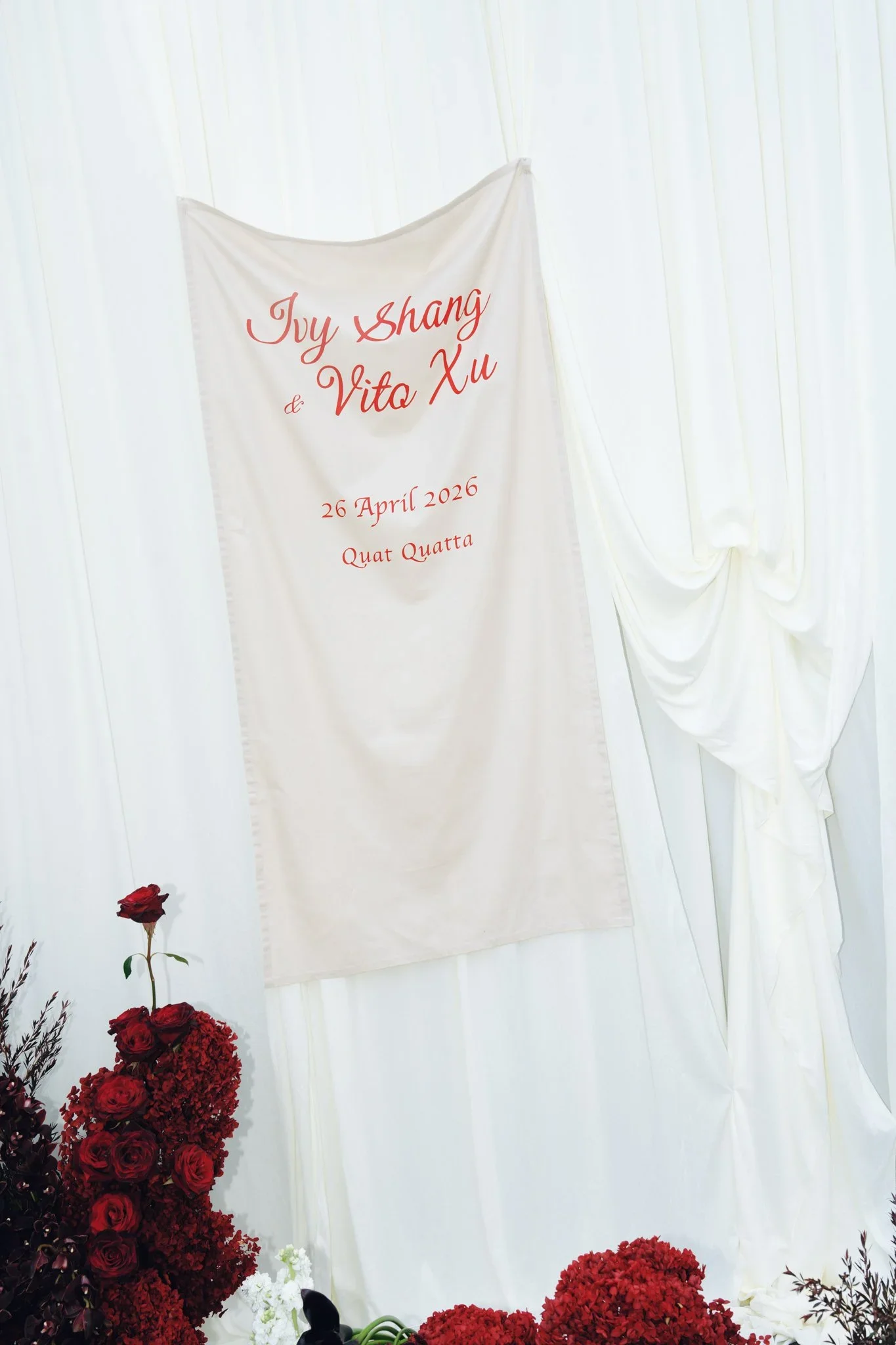 A white fabric backdrop with red cursive text reading "Juy Shang & Vita Xu  26 April 2026 Quat Quatta" and red flowers at the bottom left.