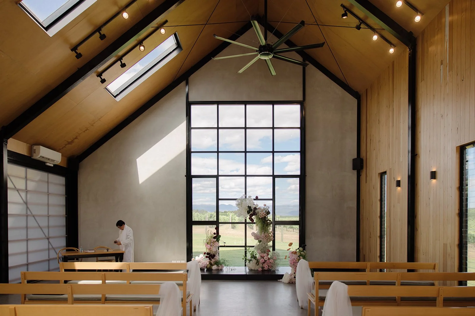 Interior of a modern chapel with wooden walls, large window, and floral arrangements at the altar. A person is standing near a desk on the left, and seats with white tissue on the backrests are arranged for visitors.