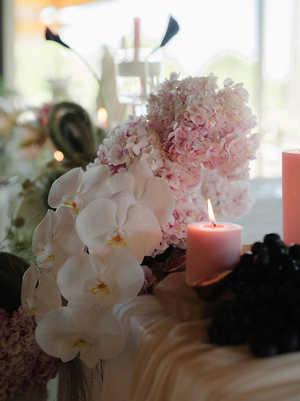 Close-up of pink hydrangeas and white orchids with candles and berries placed on a table, blurred background of windows and light.