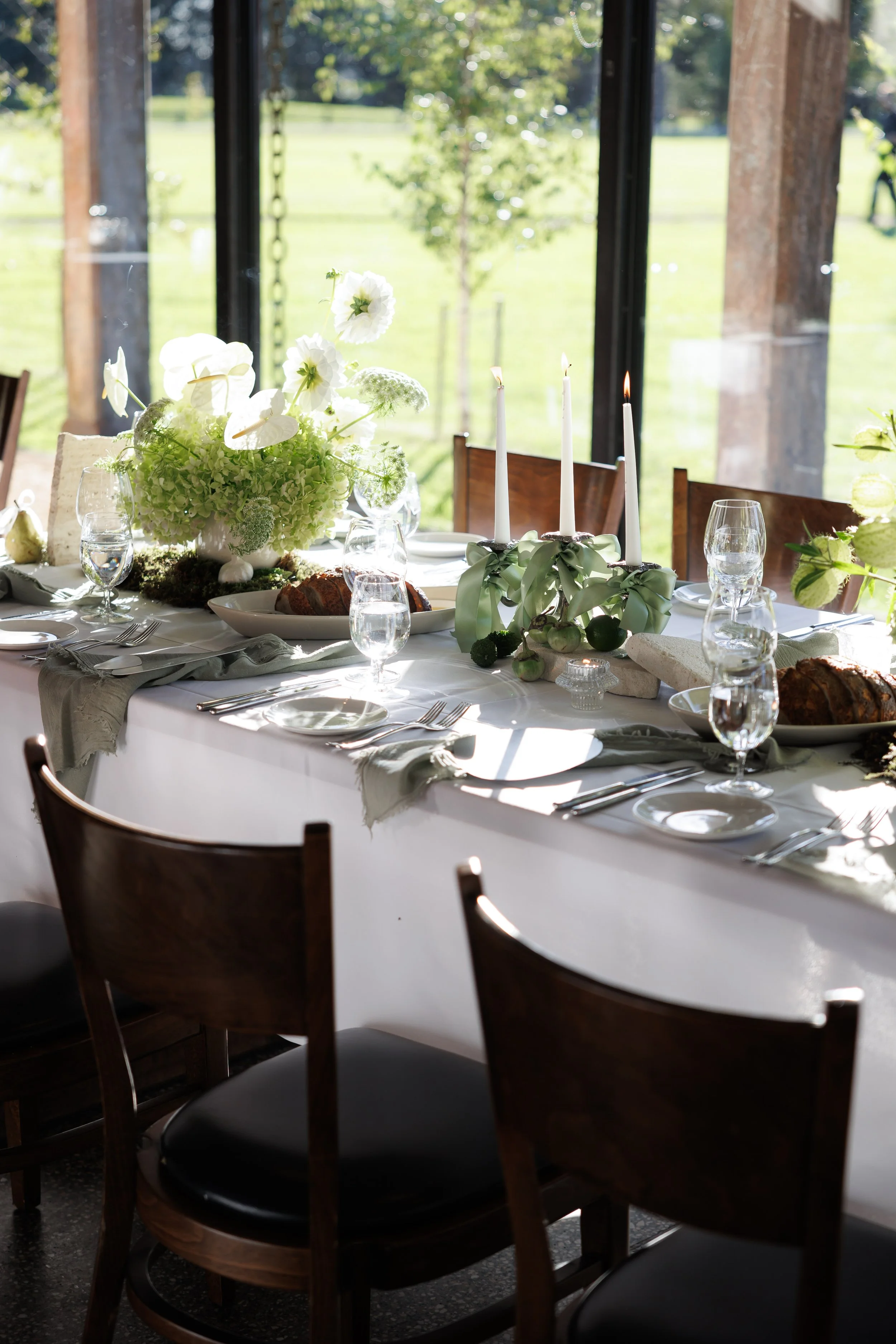 A dining table set for a meal with a large floral centerpiece, white candles, glasses, and bread, near a window with a view of a green outdoor landscape.