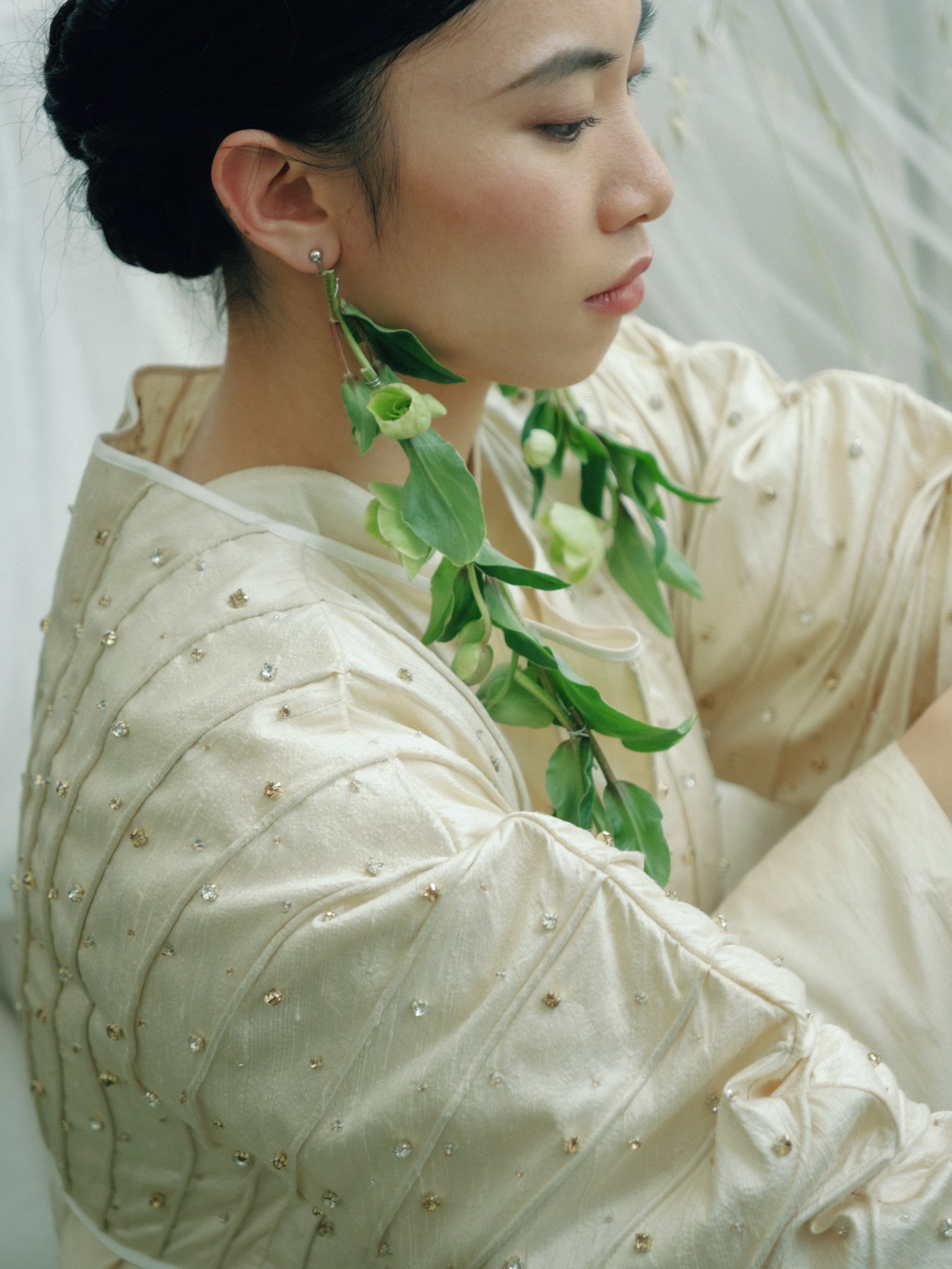 Close-up of a woman with dark hair in an updo, wearing a cream-colored, embroidered blouse with pearl details, and accessorized with leaf-shaped earrings and a matching leafy necklace.