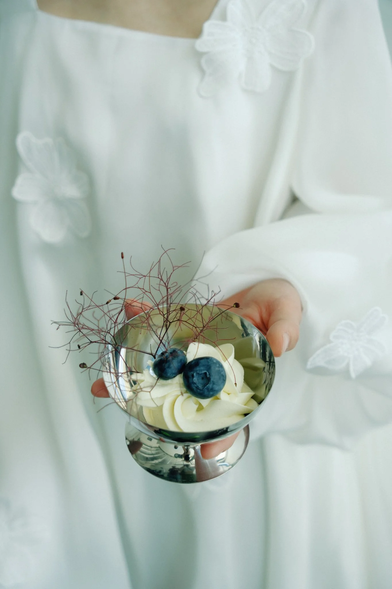 A person holding a glass bowl of blueberries and whipped cream, decorated with edible garnishes and sugar spun threads, in a white dress with floral embroidery.