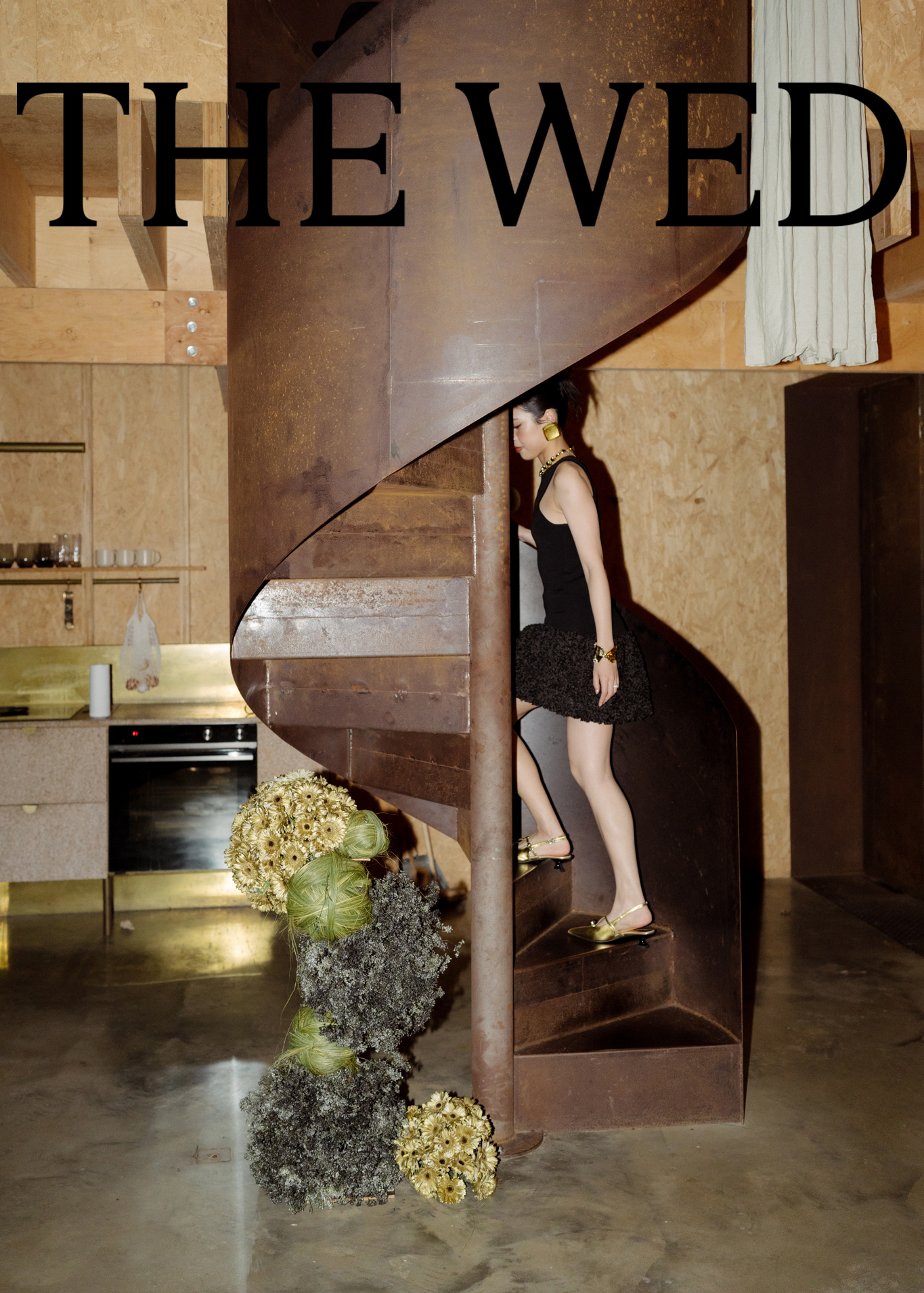 A woman in a black dress and gold shoes is walking down a spiral staircase in a rustic interior with wooden walls. The staircase is made of rust-colored metal. There is a large bouquet of flowers at the bottom of the staircase.