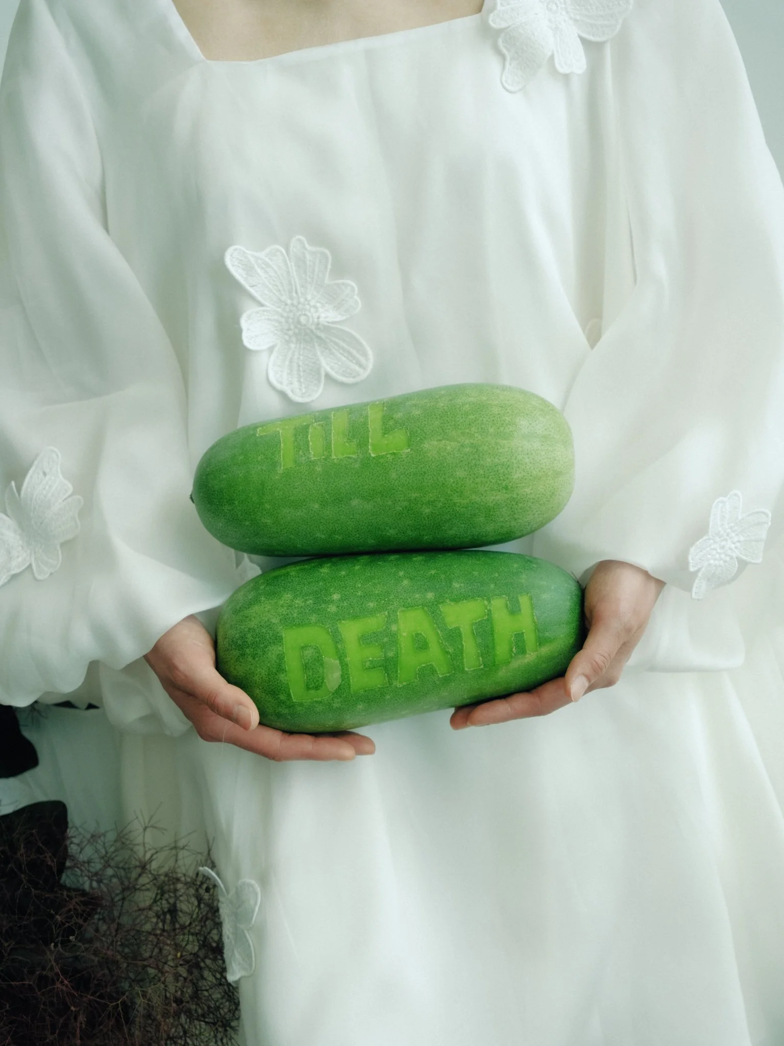 A person holding two cucumbers, one with the word 'DEATH' carved into its skin, wearing a white embroidered dress.