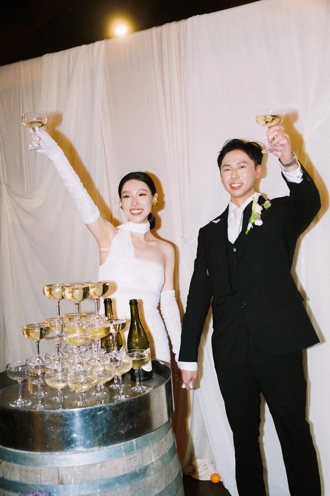A bride and groom at their wedding celebration raise glasses of champagne for a toast, standing next to a champagne tower, with a white draped wall in the background.