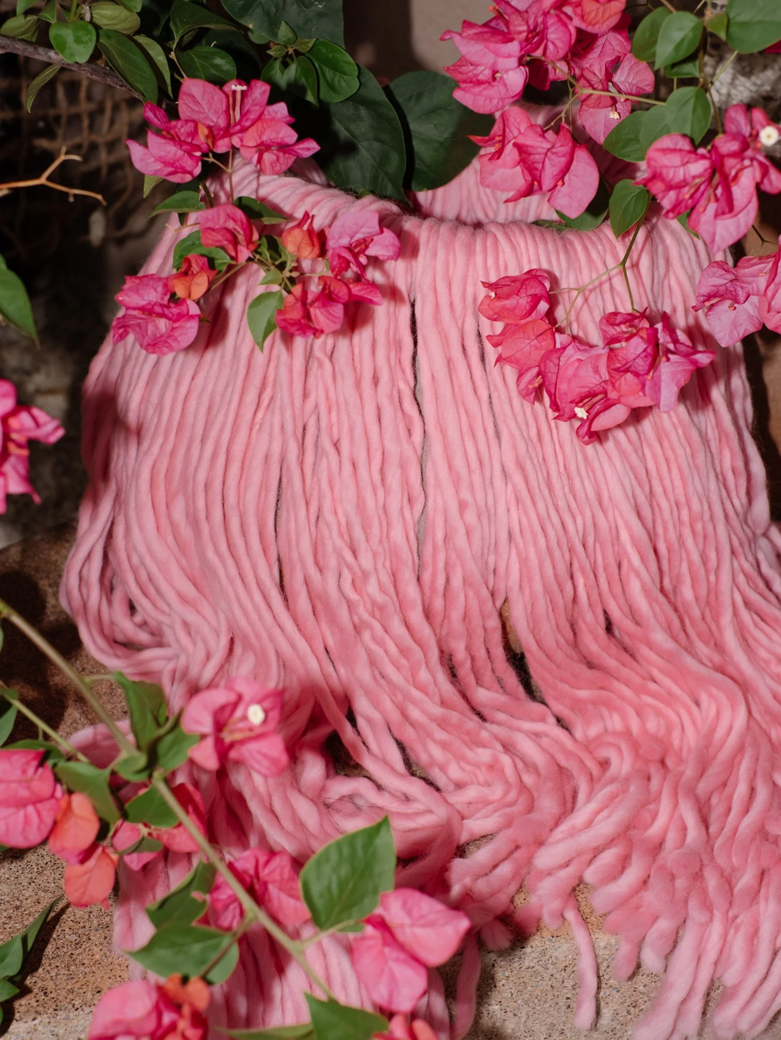 Pink crocheted blanket surrounded by pink bougainvillea flowers and green leaves.