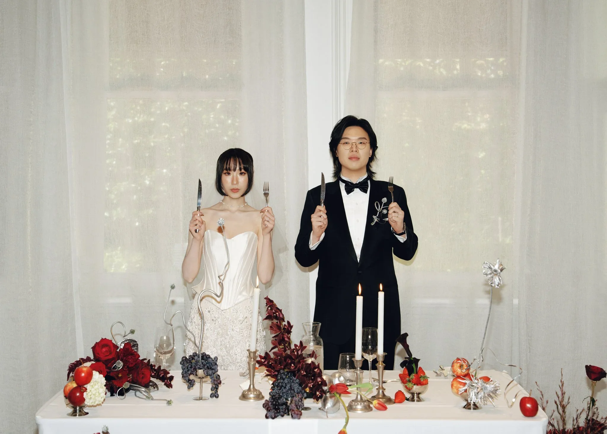 A bride and groom standing behind a decorated table with candles and fruit, holding cutlery, in front of a white curtain.