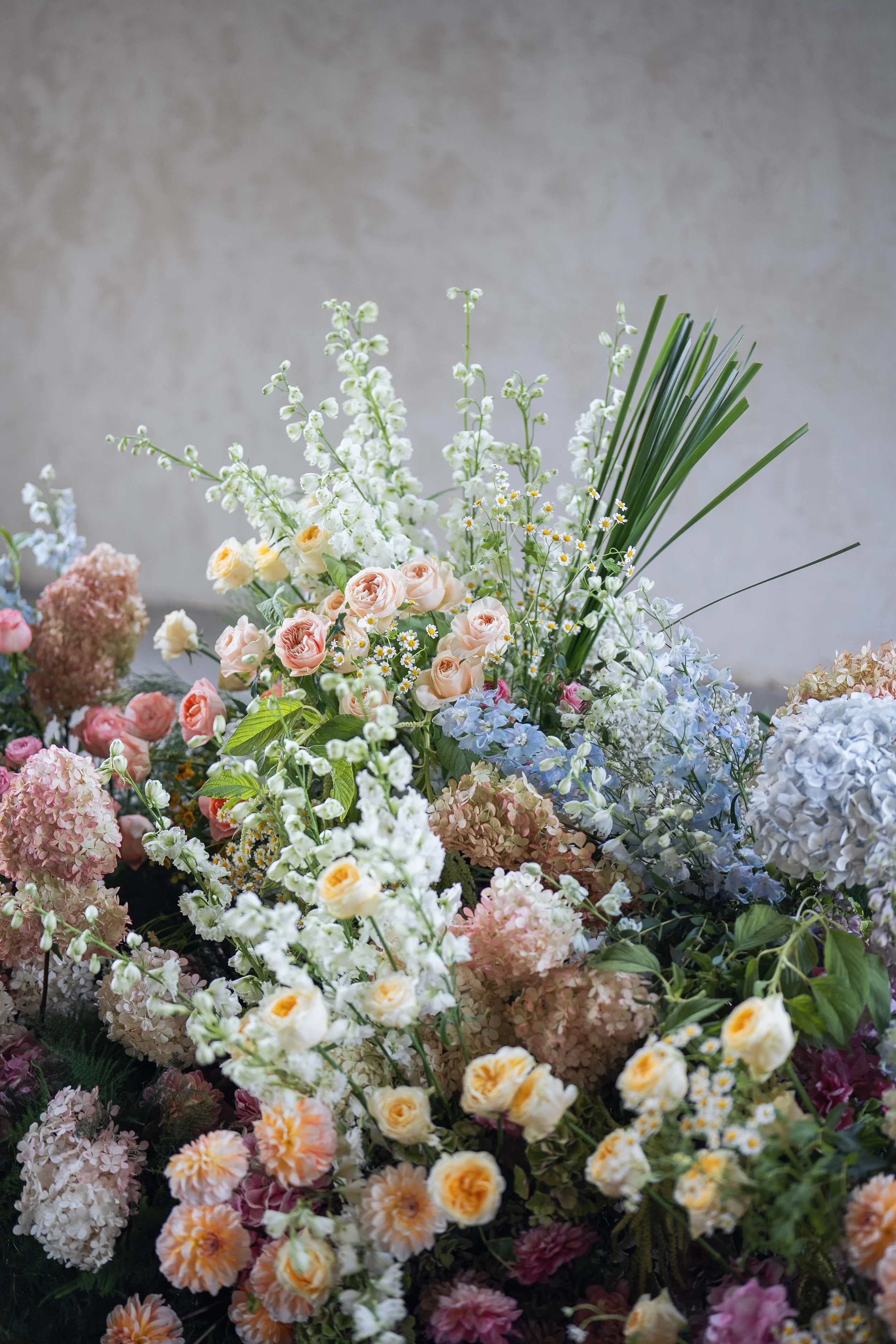 A bouquet of mixed flowers including white, pink, peach, and purple blossoms with green leaves and grass-like foliage against a neutral background.