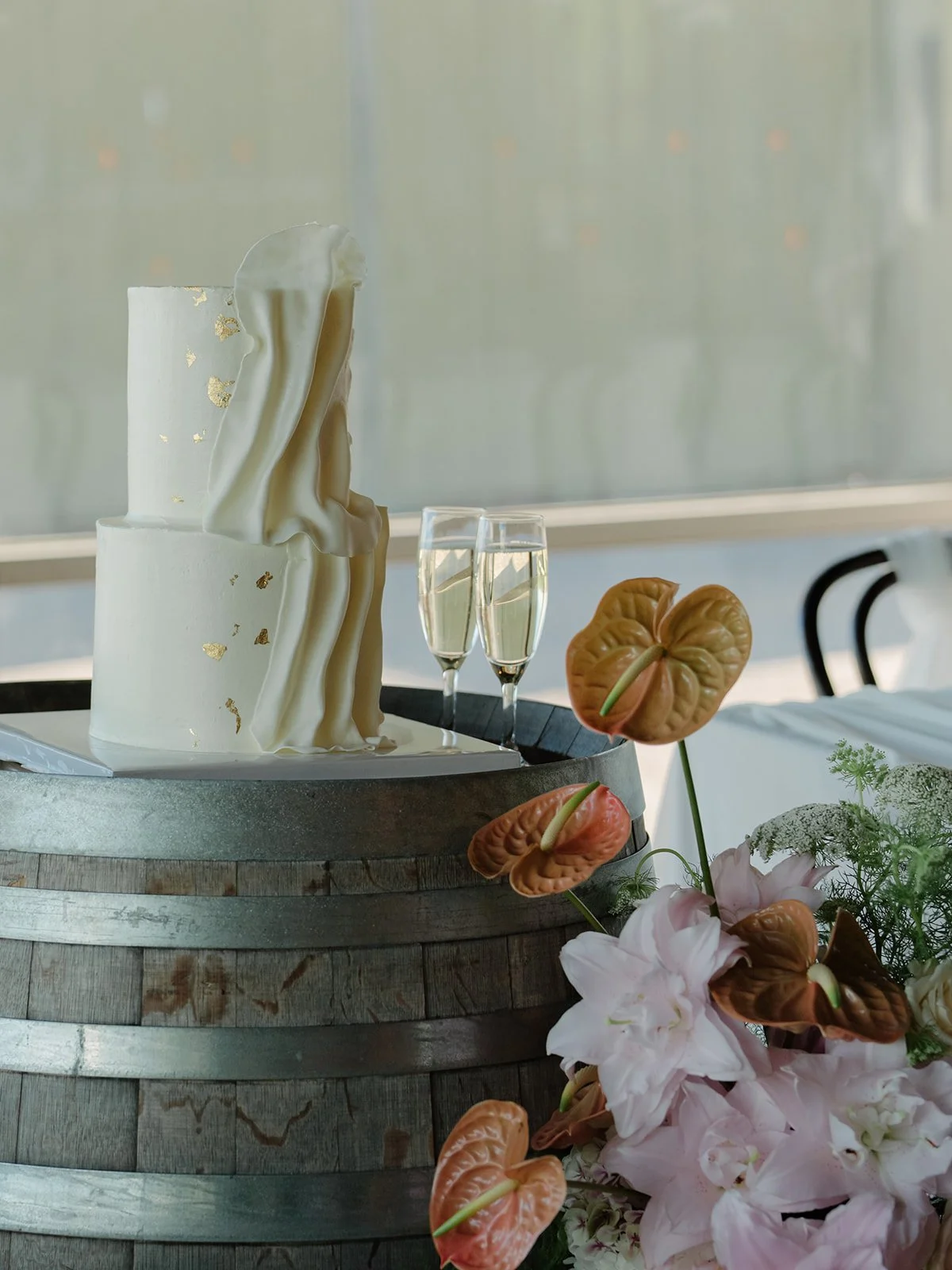A two-tiered white cake with gold leaf accents and ruffled fondant draping, placed on a wooden barrel, with two glasses of champagne, orange flowers, and pink flowers nearby, set in a bright room with a large window.