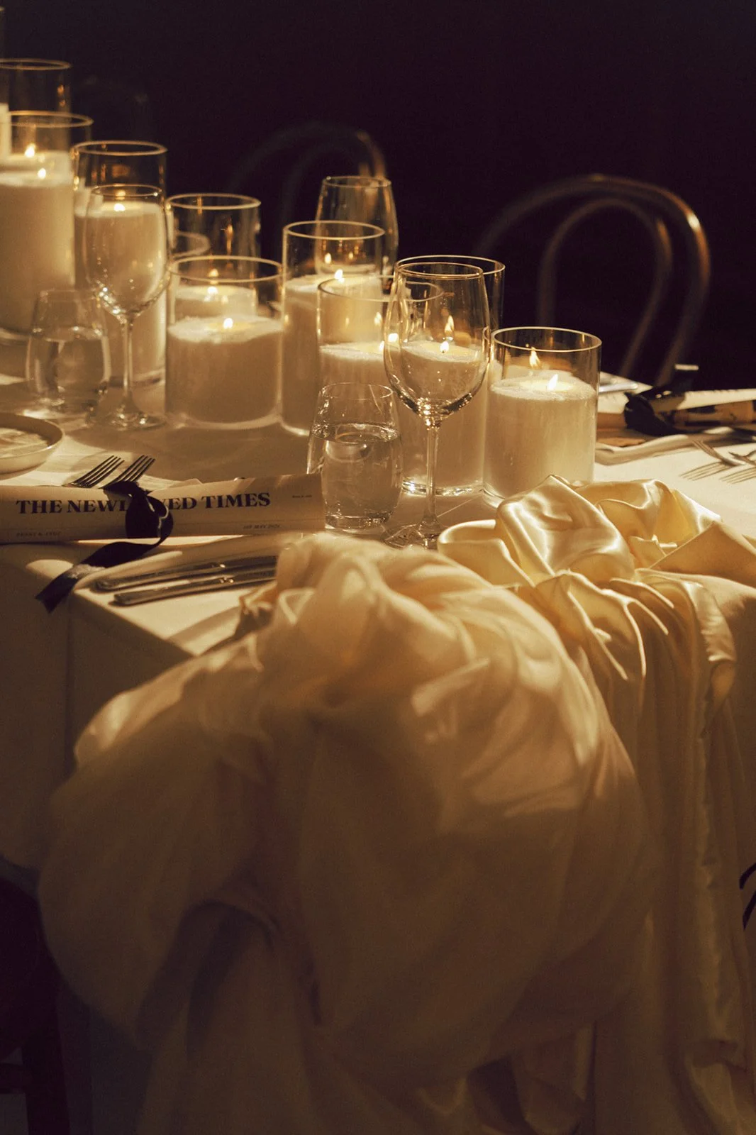 A formal dining table set with lit candles in glass holders, wine glasses, silverware, a rolled newspaper, and elegant cream-colored cloth napkins or dressings.
