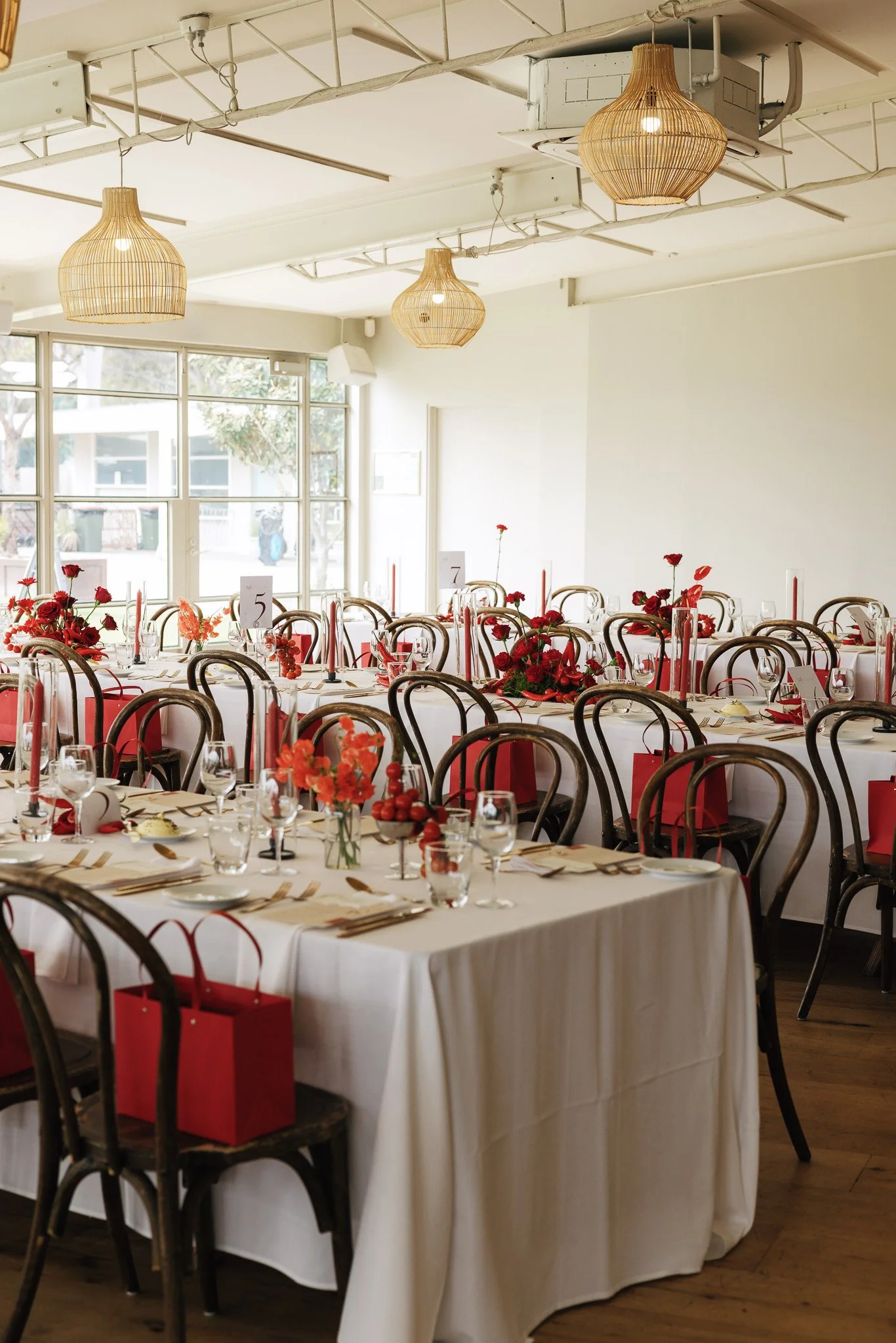 Elegant banquet hall set for an event with round tables covered in white tablecloths, decorated with red flowers, candles, and glassware. Wooden chairs surround the tables, and red gift bags are placed on or near the chairs. Large windows let in natu