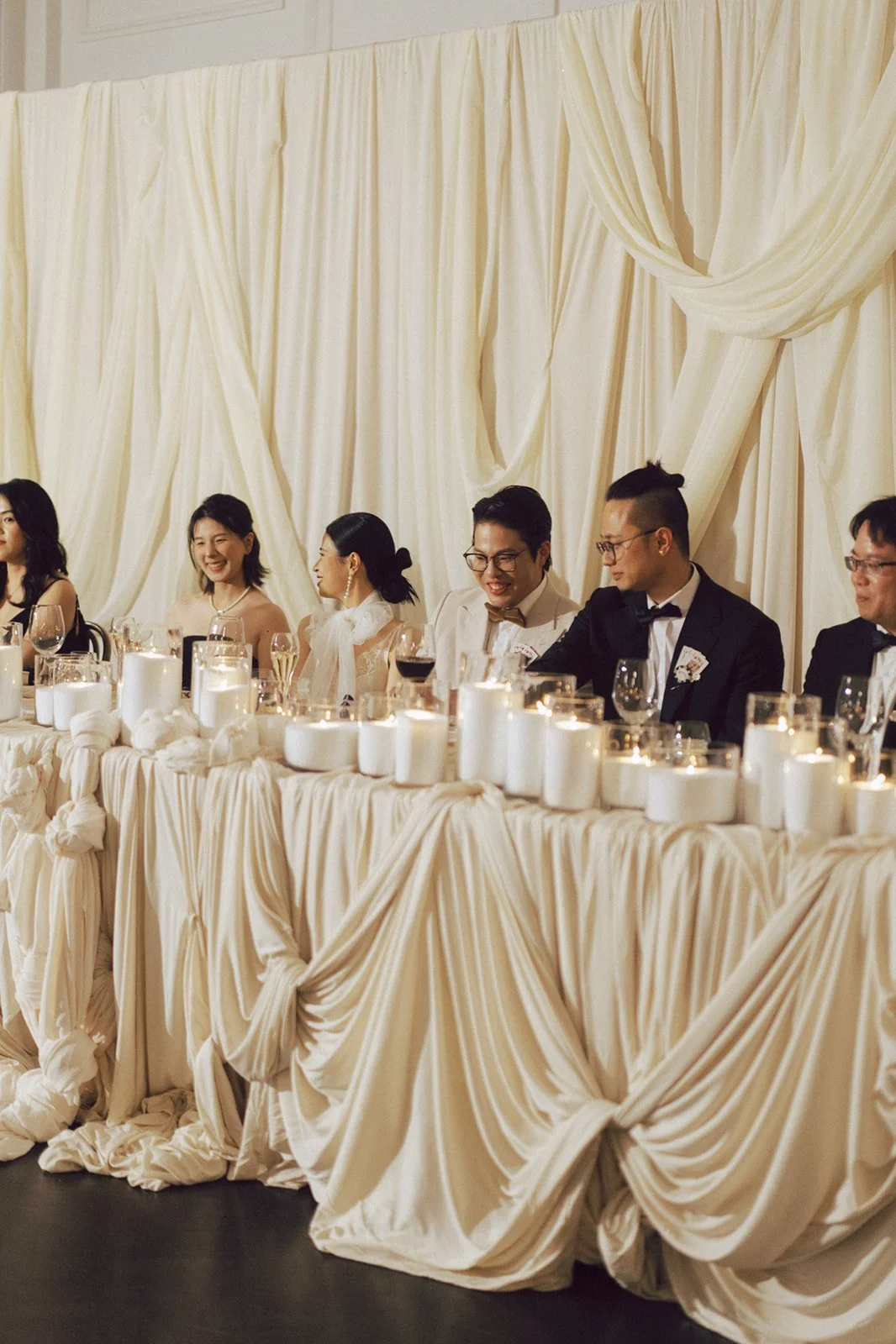 Group of Asian people dressed in formal attire at a wedding reception table decorated with candles and draped fabric decor.