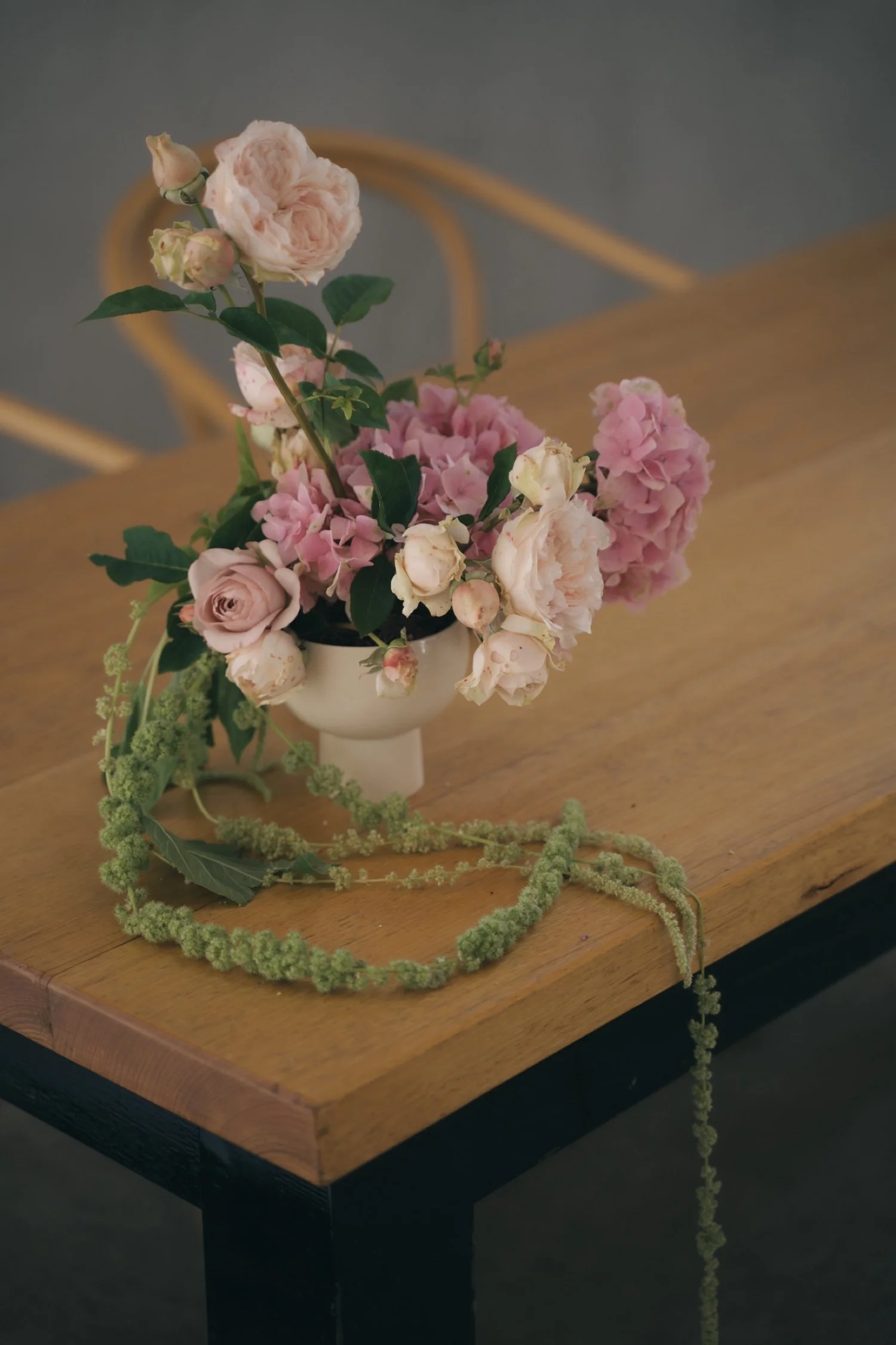 A bouquet of pink and cream roses and hydrangeas in a white vase, placed on a wooden table with a decorative green trailing plant around the base.