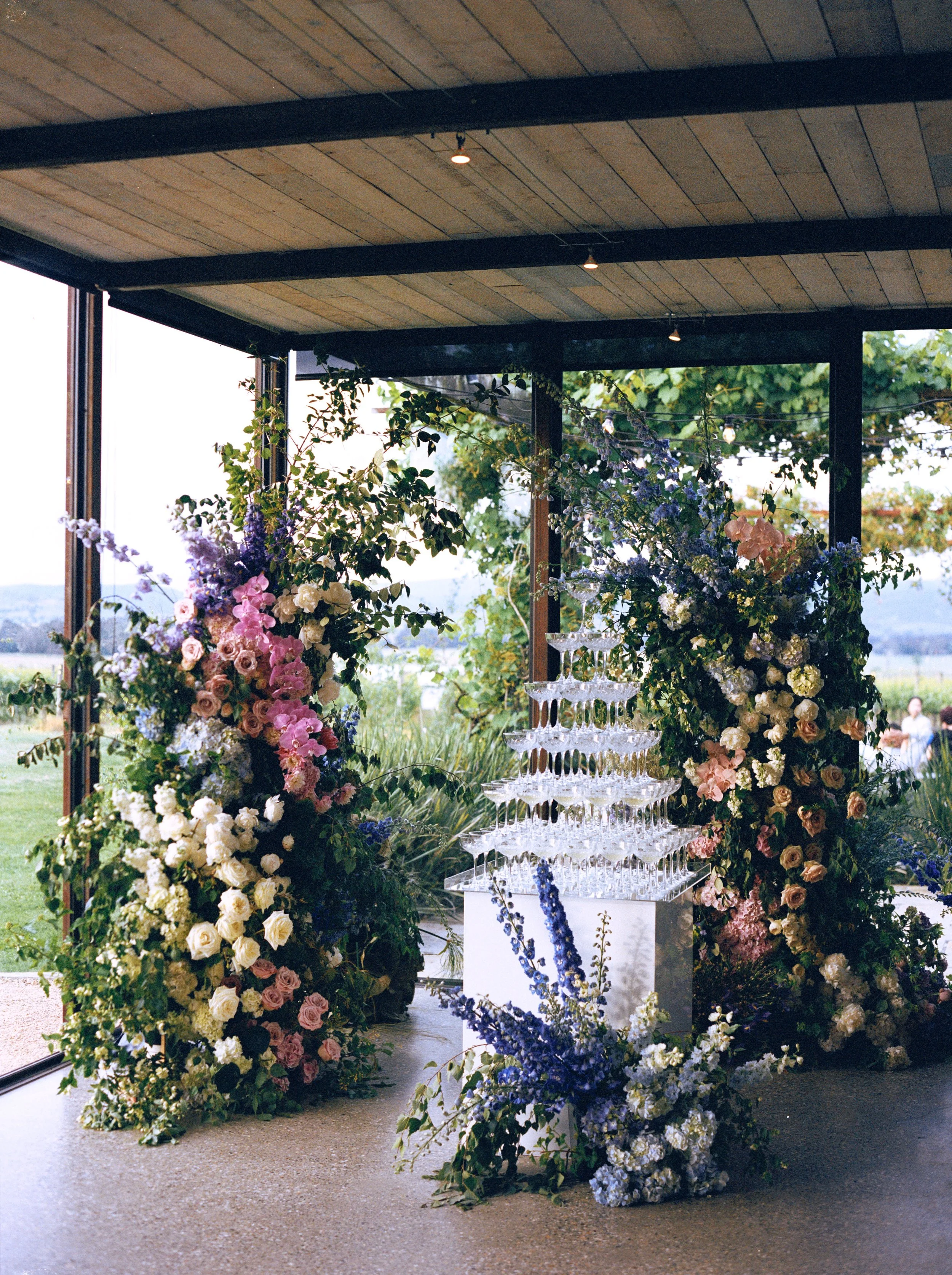 A floral arrangement with pink, white, and purple flowers and lush greenery surrounds a pyramid of clear glasses, set up indoors with a wooden ceiling and large windows showing an outdoor landscape.