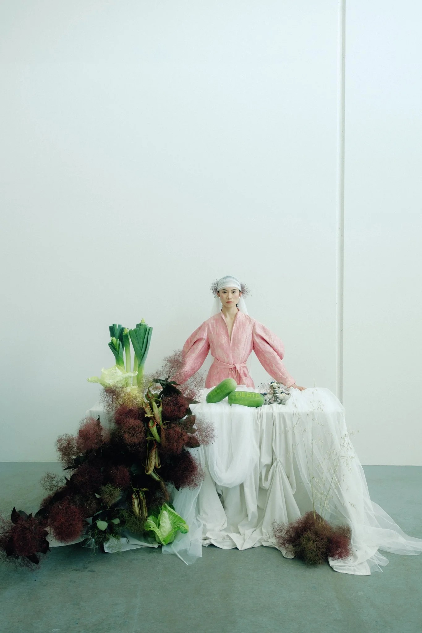 A woman wearing a pink traditional dress and head covering stands behind a table decorated with various vegetables and plants, in a plain white-walled room.