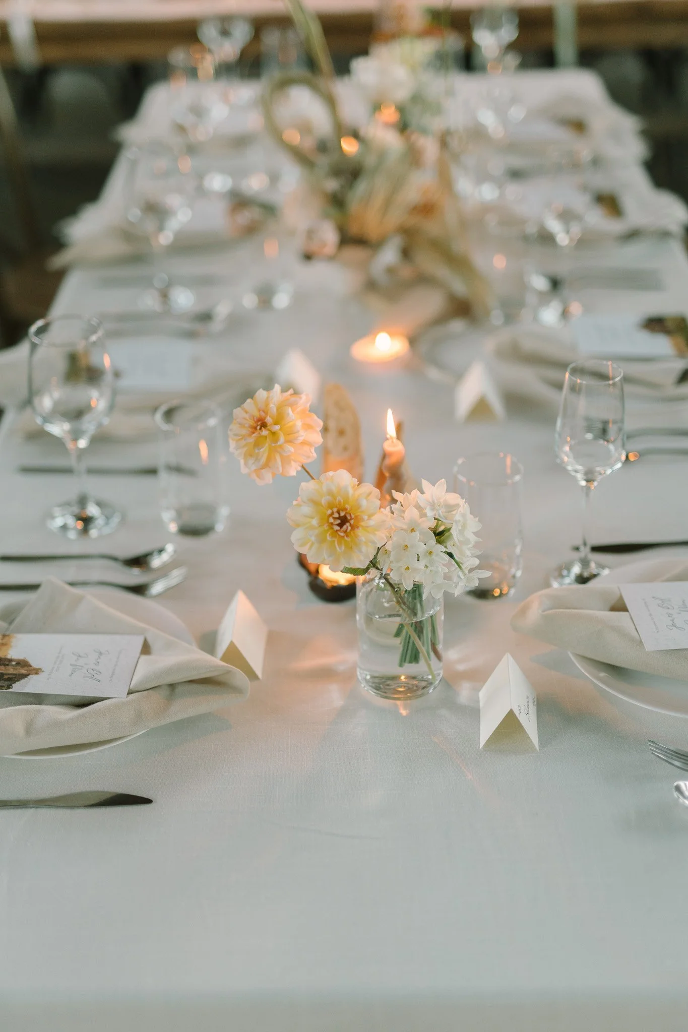 A wedding or formal event table setting with white tablecloth, floral centerpieces, candles, clear glassware, napkins, and printed menus or place cards, in a well-lit indoor space.