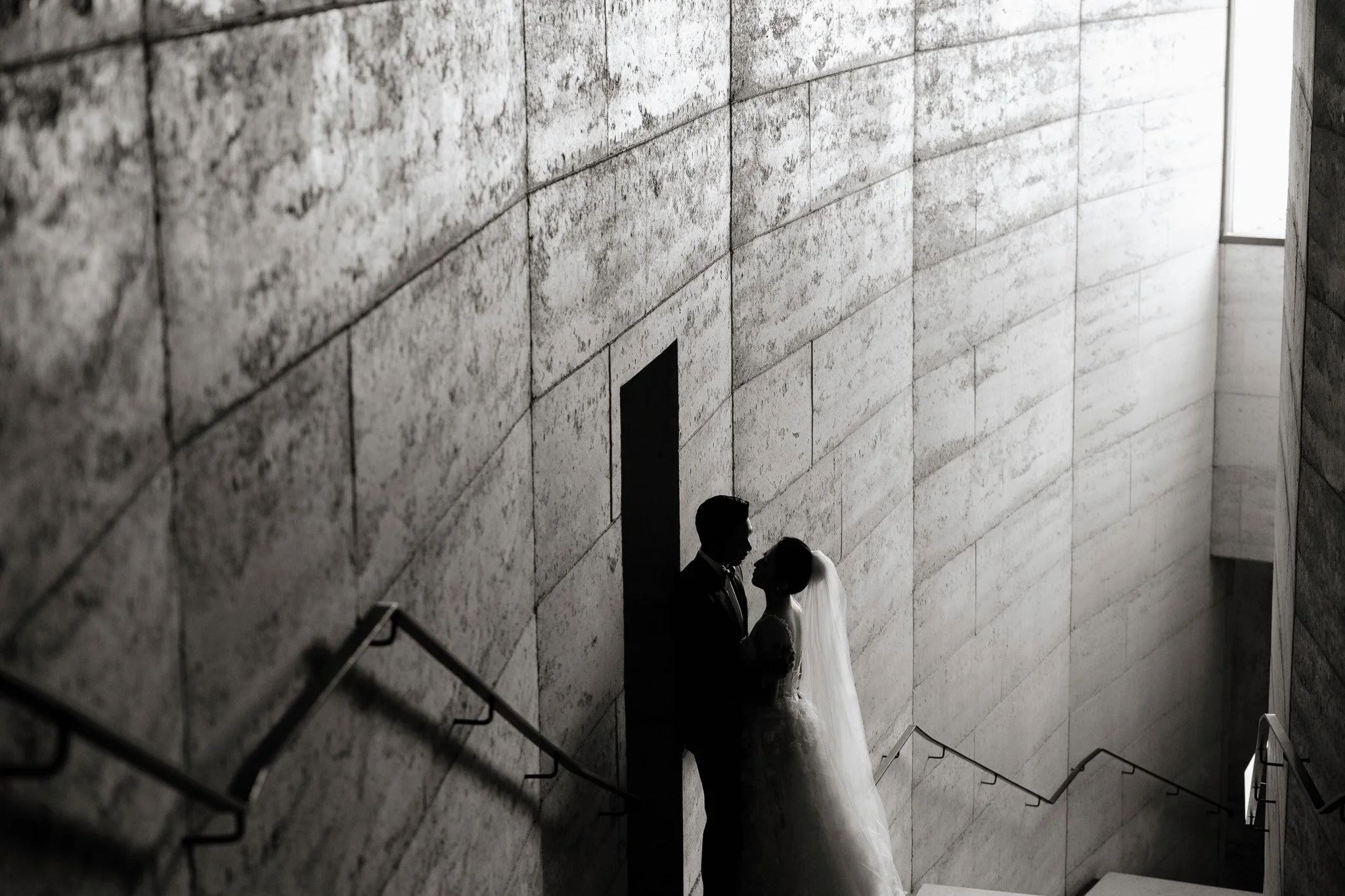 A bride and groom standing close together at the bottom of a staircase in a modern building with a textured stone wall and large windows.