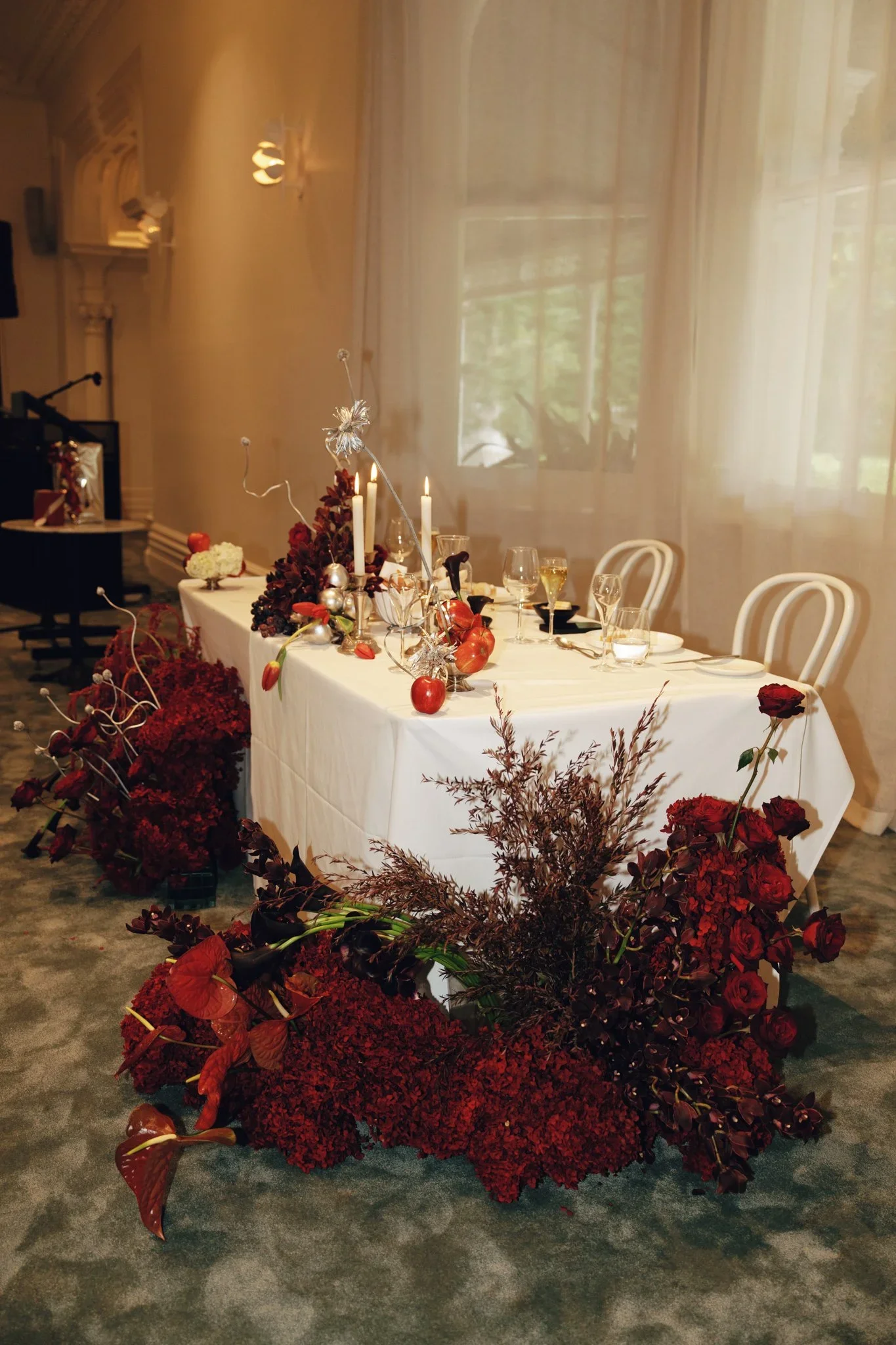 A long dining table decorated with red and white floral arrangements, candles, and glassware, surrounded by white chairs, in a warmly lit room with large windows and sheer curtains.