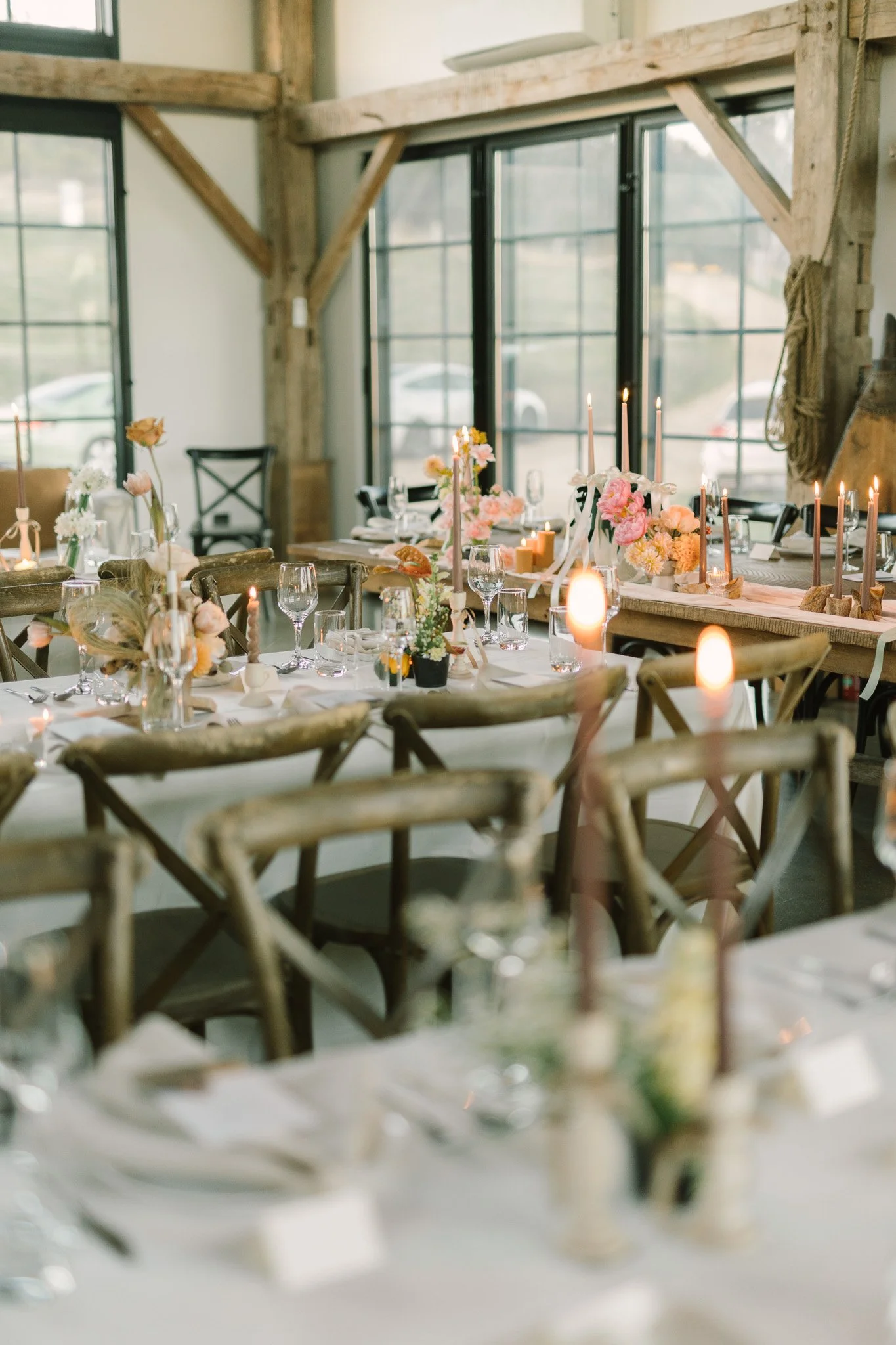 Wedding reception table decorated with pink flowers, candles, and glassware inside a rustic venue with large windows and wooden beams.