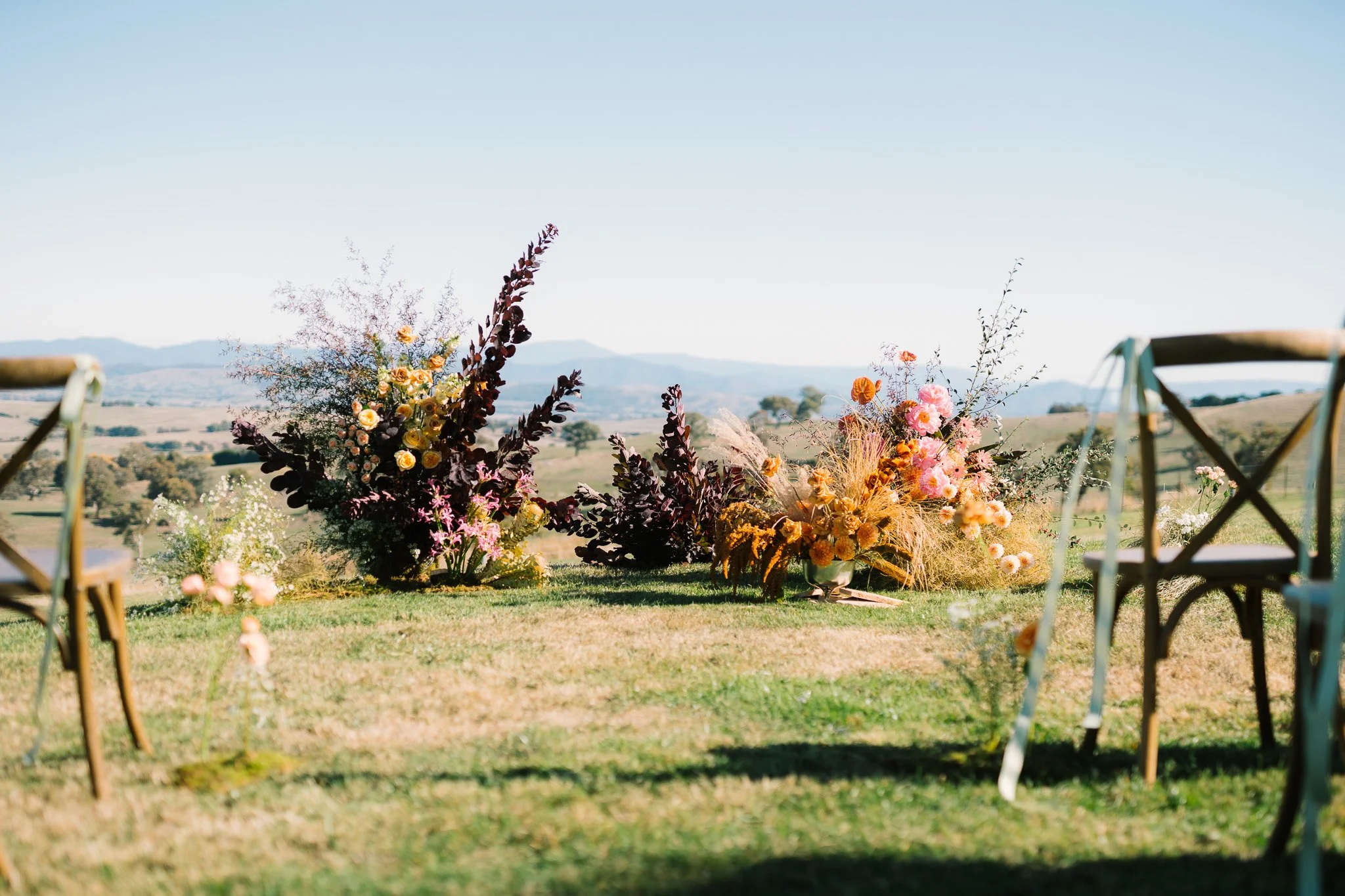 Outdoor wedding reception setup with a floral arrangement on the ground and two chairs facing it, with a scenic landscape of mountains and fields in the background.