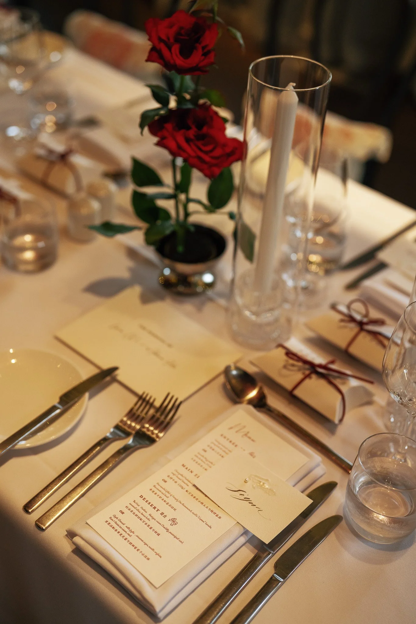 A dining table setup with a small potted red rose as a centerpiece, a tall glass candle holder with a white candle, a menu, silverware, and water glasses. The table is elegantly arranged, likely for a formal event or dinner.