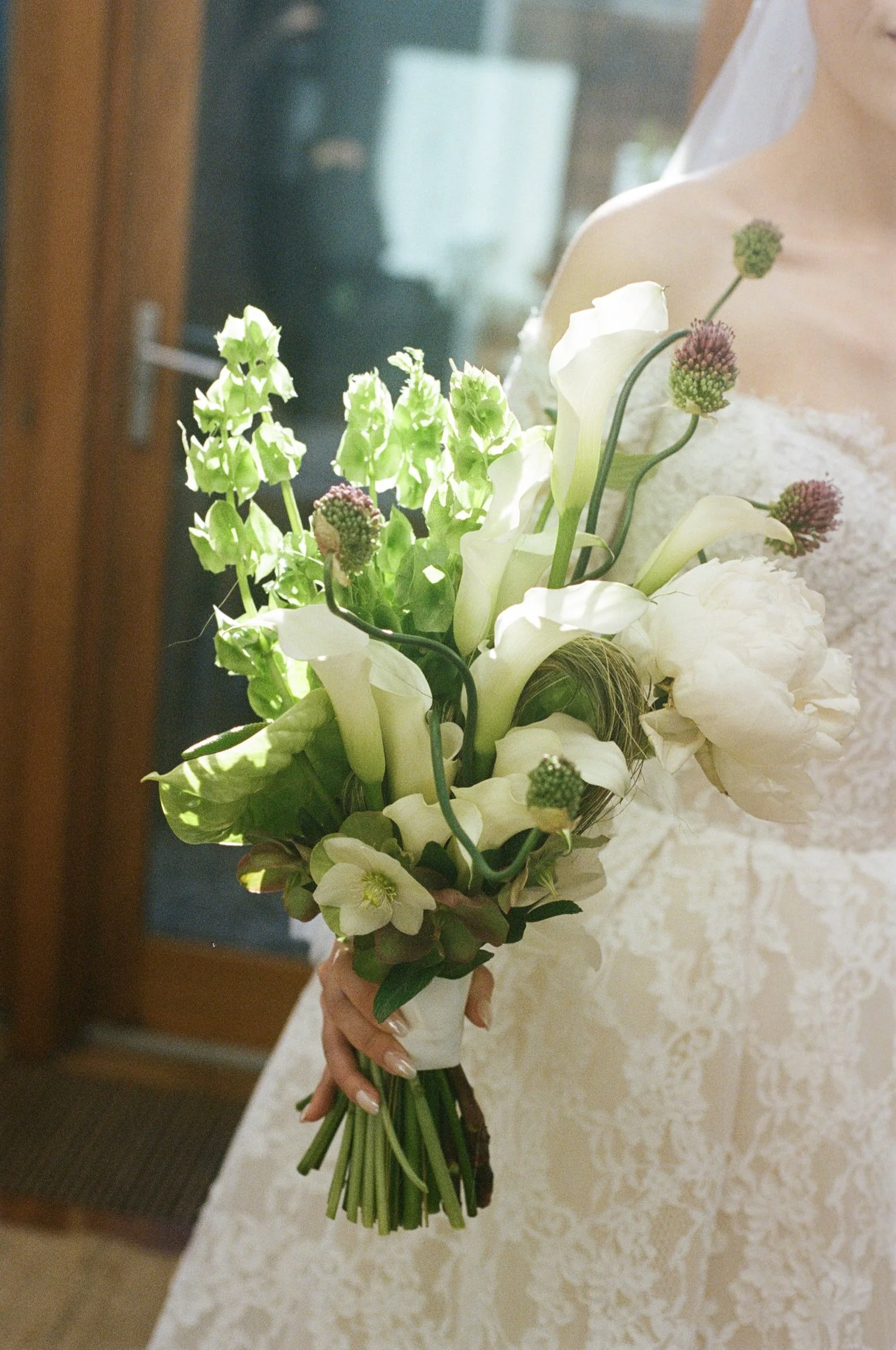 A bride holding a bouquet of white flowers and greenery, including calla lilies and other blooms, near a window with a lace wedding dress.