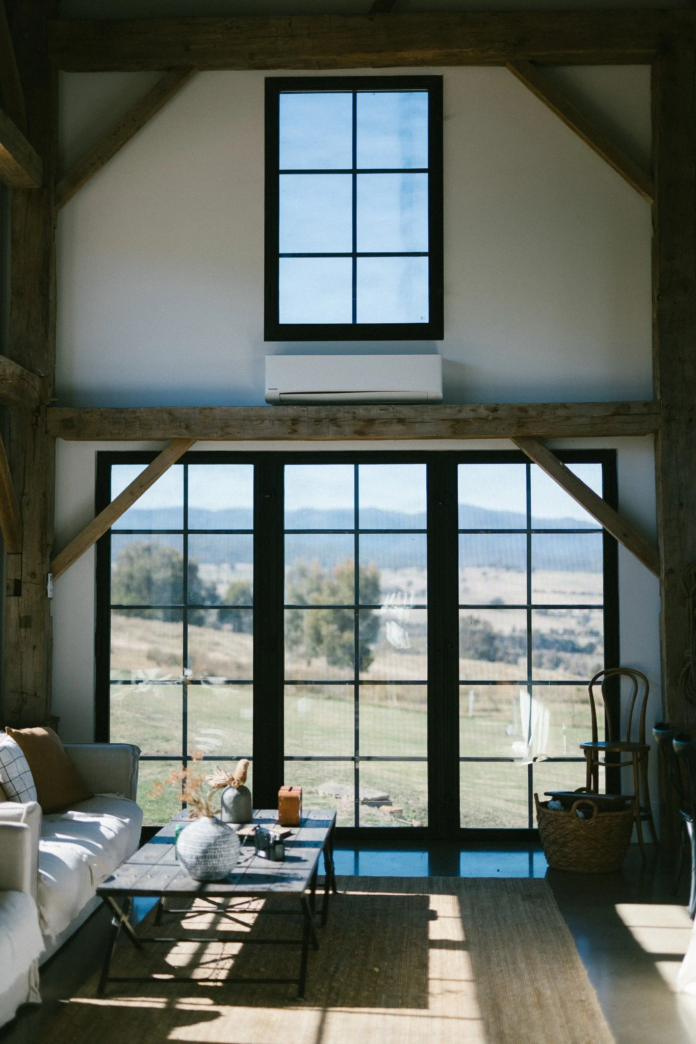 Living room with large black-framed windows and door, white walls, wooden beams, and furniture including a white sofa, wooden coffee table, chair, and basket, with a view of open countryside outside.