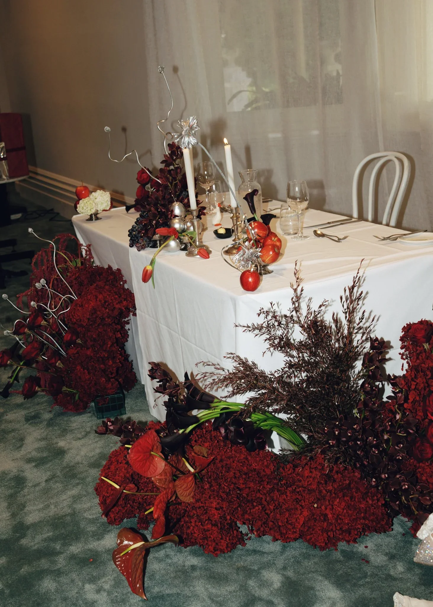 A decorated dining table with floral arrangements, candles, and tableware surrounded by red and dark purple flowers.