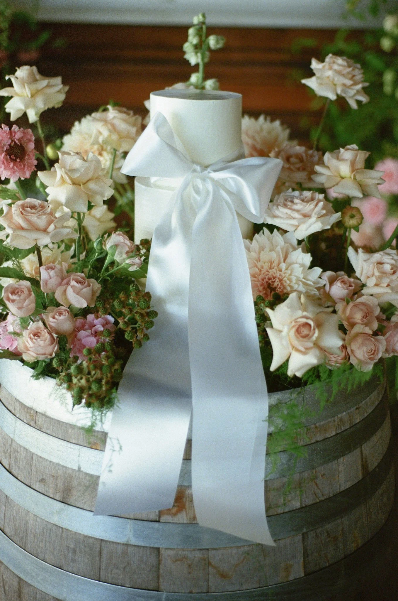 A flower arrangement with pink and white roses, white and green flowers, and greenery, placed on a wooden, circular table with a white ribbon tied in a bow around a cylindrical object in the center.