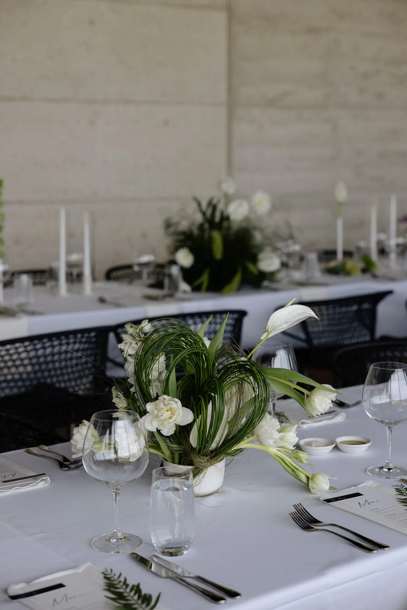 Elegant banquet table with white tablecloth, floral centerpiece with white flowers and greenery, wine glasses, water glasses, silverware, and place cards in a modern venue with a concrete wall background.