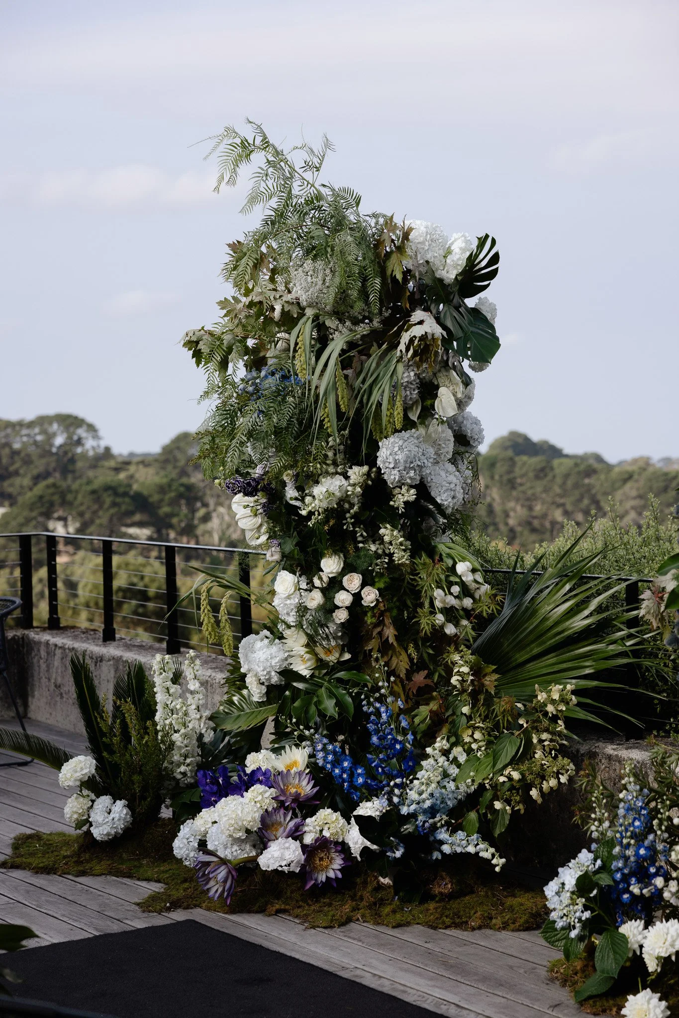 A large arrangement of mostly white flowers with some purple and blue accents, along with green foliage, set on an outdoor deck with a forested landscape in the background.