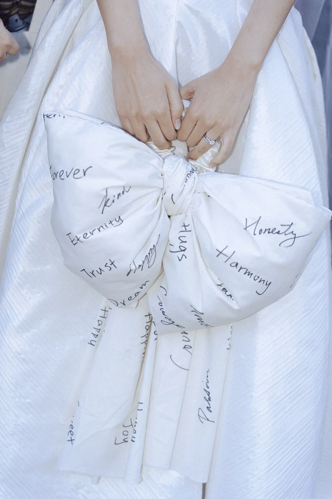 Close-up of a bride holding her hands in front of her wedding dress, which has handwritten words like 'Trust,' 'Eternity,' 'Honesty,' and 'Harmony' on the fabric.