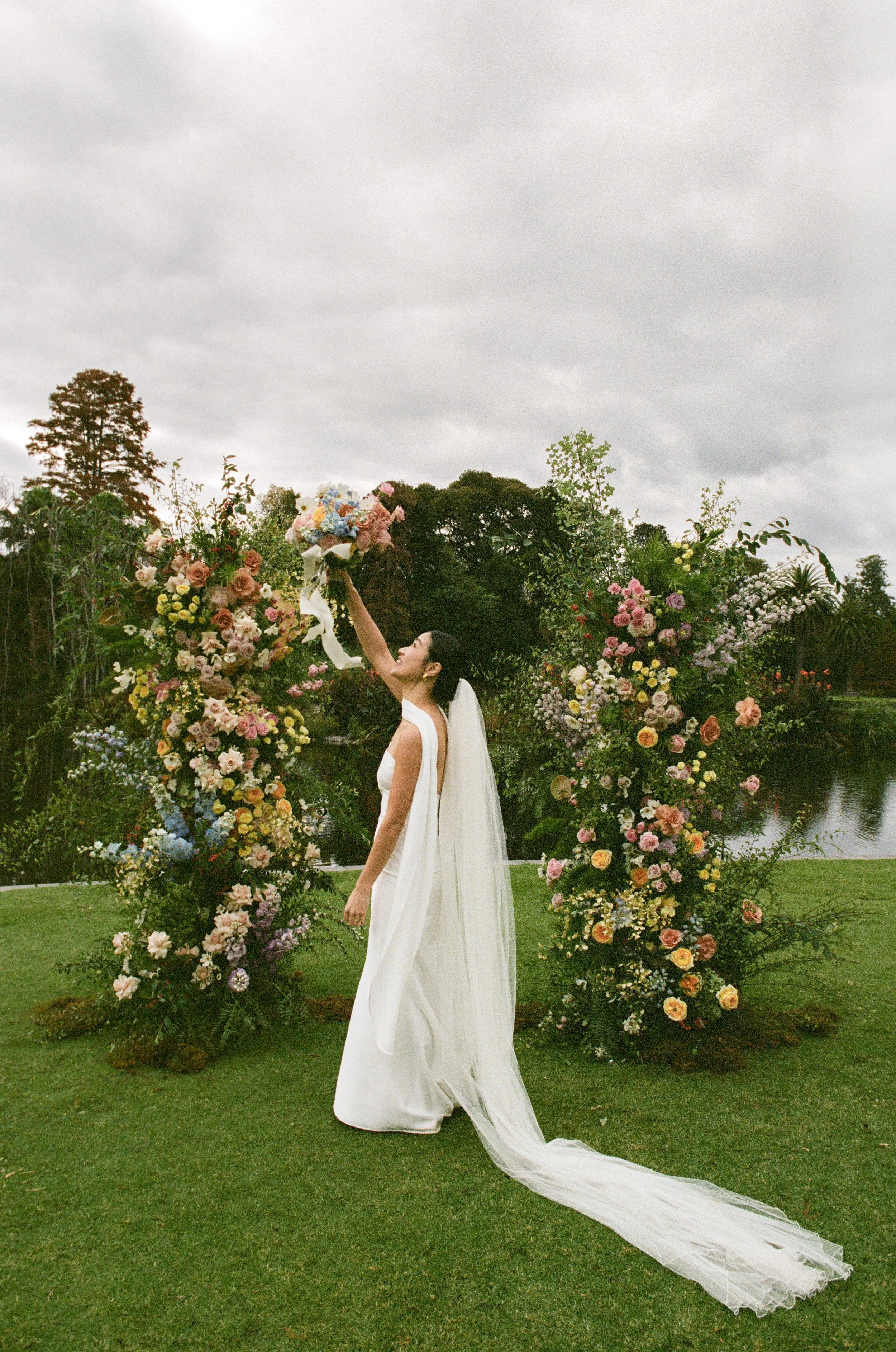 A bride in a wedding dress holding a bouquet of flowers, standing in front of a floral arch outdoors on a cloudy day.