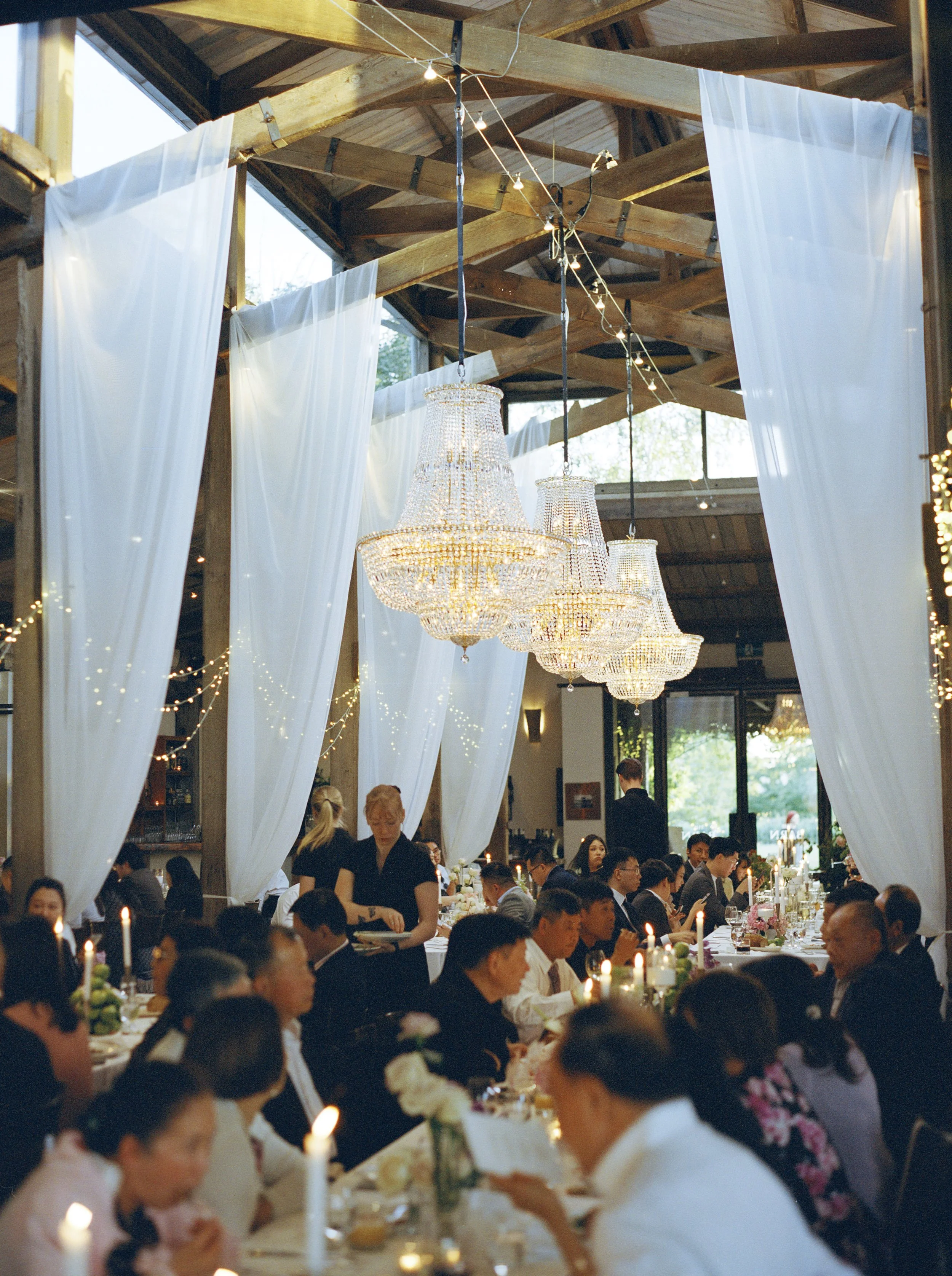 Guests seated at banquet tables during a wedding reception or event inside a rustic venue with wooden beams and chandeliers, surrounded by white draped curtains and string lights.