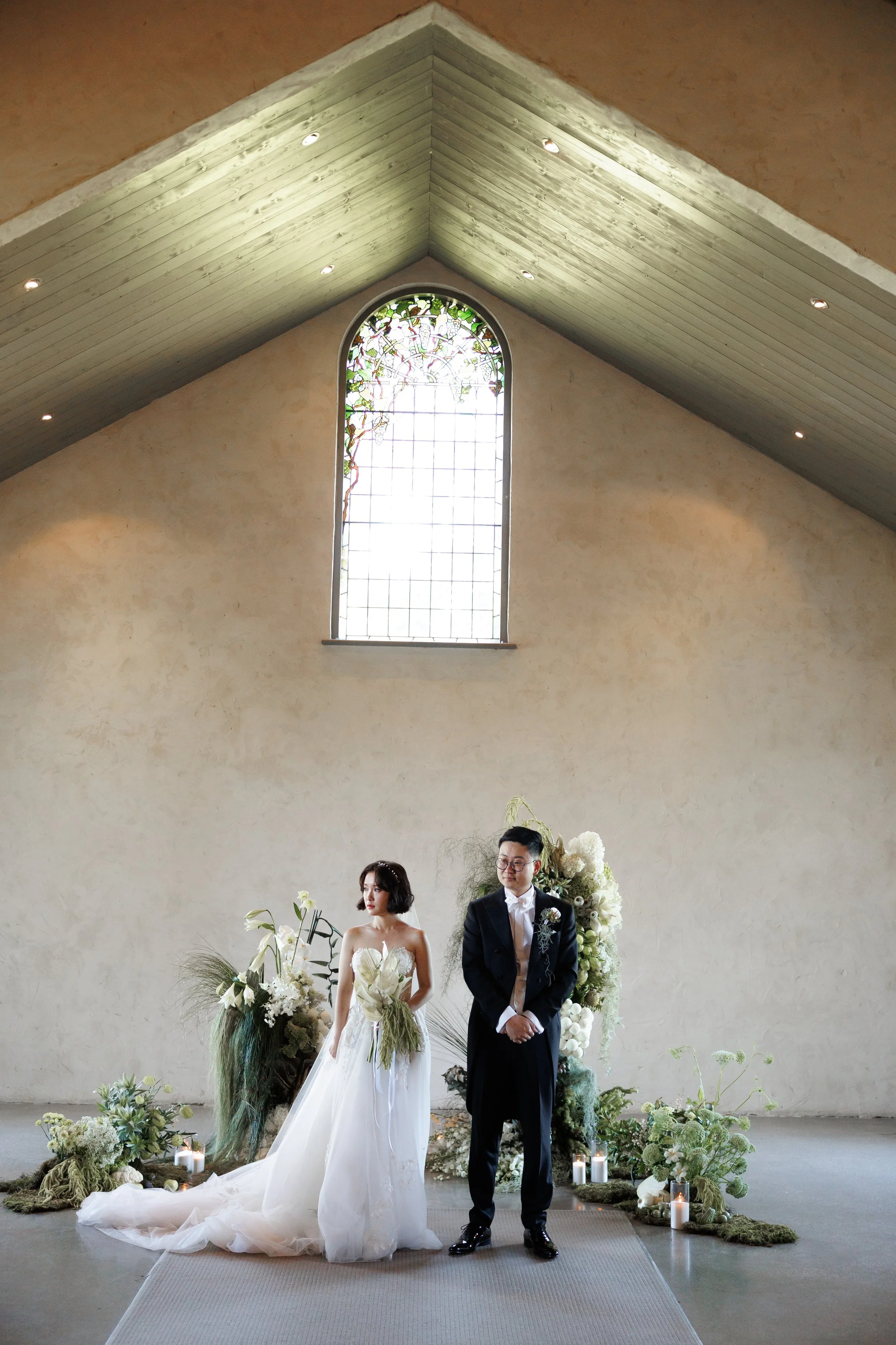 A bride and groom standing at the altar during their wedding inside a church with a large stained glass window, surrounded by white flowers, candles, and greenery.