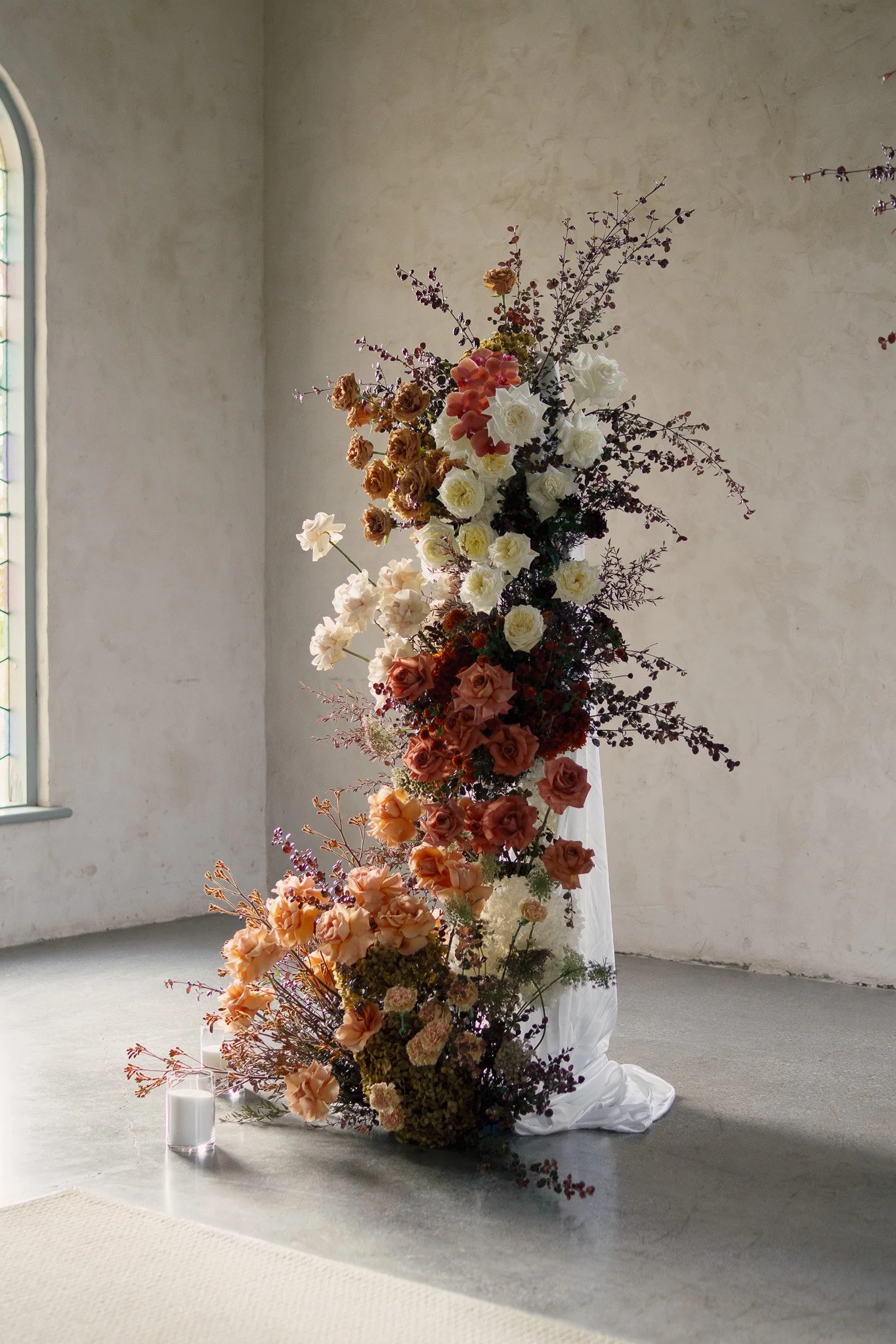 A tall, elaborate floral arrangement featuring various flowers in shades of peach, white, and deep red, placed inside a church with a minimalist design. The flowers cascade downwards on a white fabric-draped stand, with soft natural light coming thro