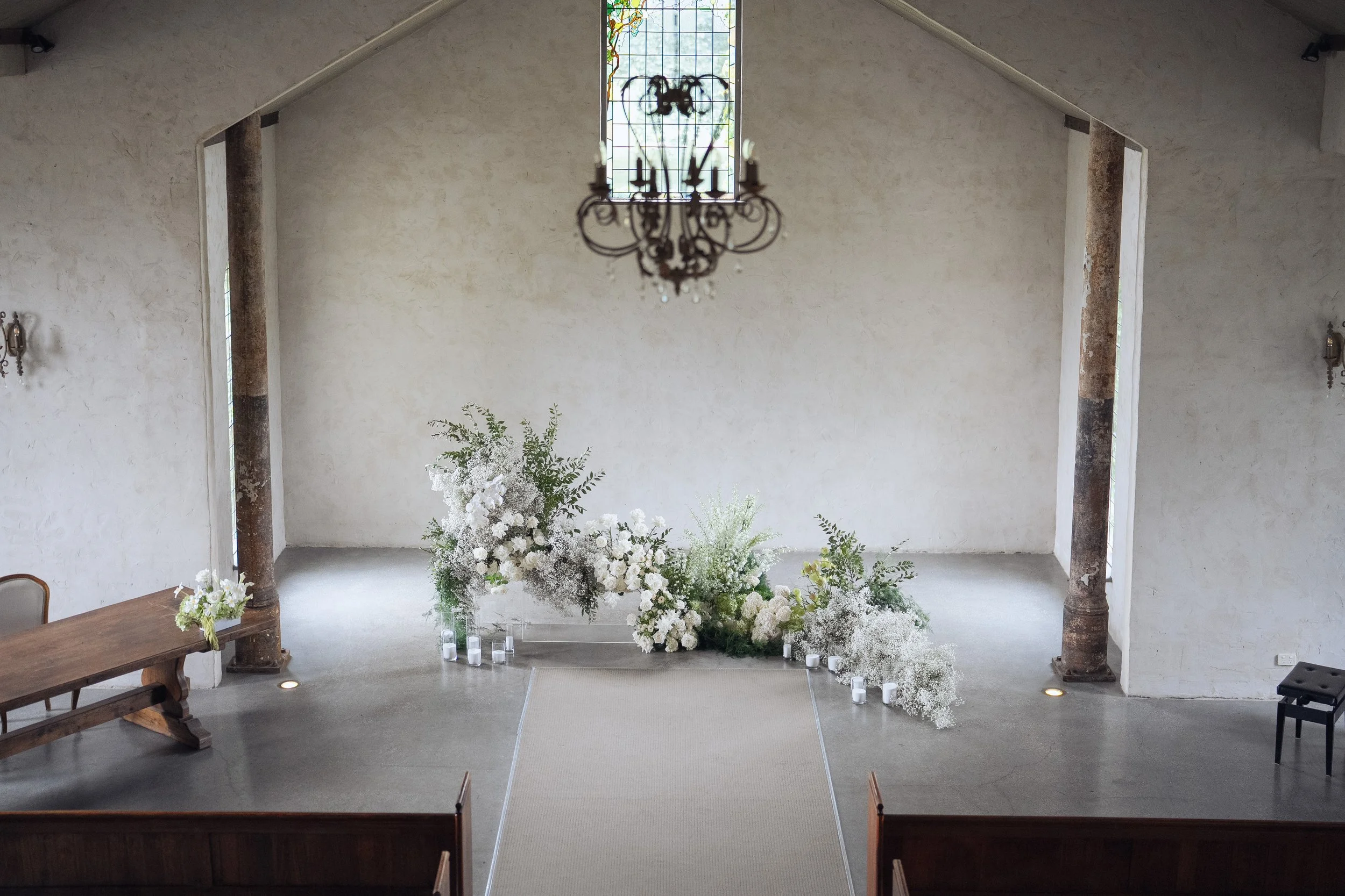 Interior of a church or wedding venue with a high ceiling, a stained glass window, a chandelier, and floral arrangements on the floor, likely set up for a wedding ceremony.