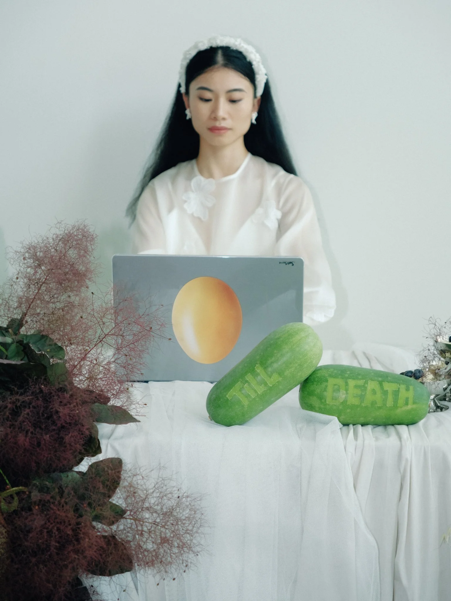 A woman with long dark hair, dressed in a white outfit with floral details, sitting behind a computer. There are decorative plants and large green stones with the words 'TIME' and 'DEATH' carved into them on a white table.