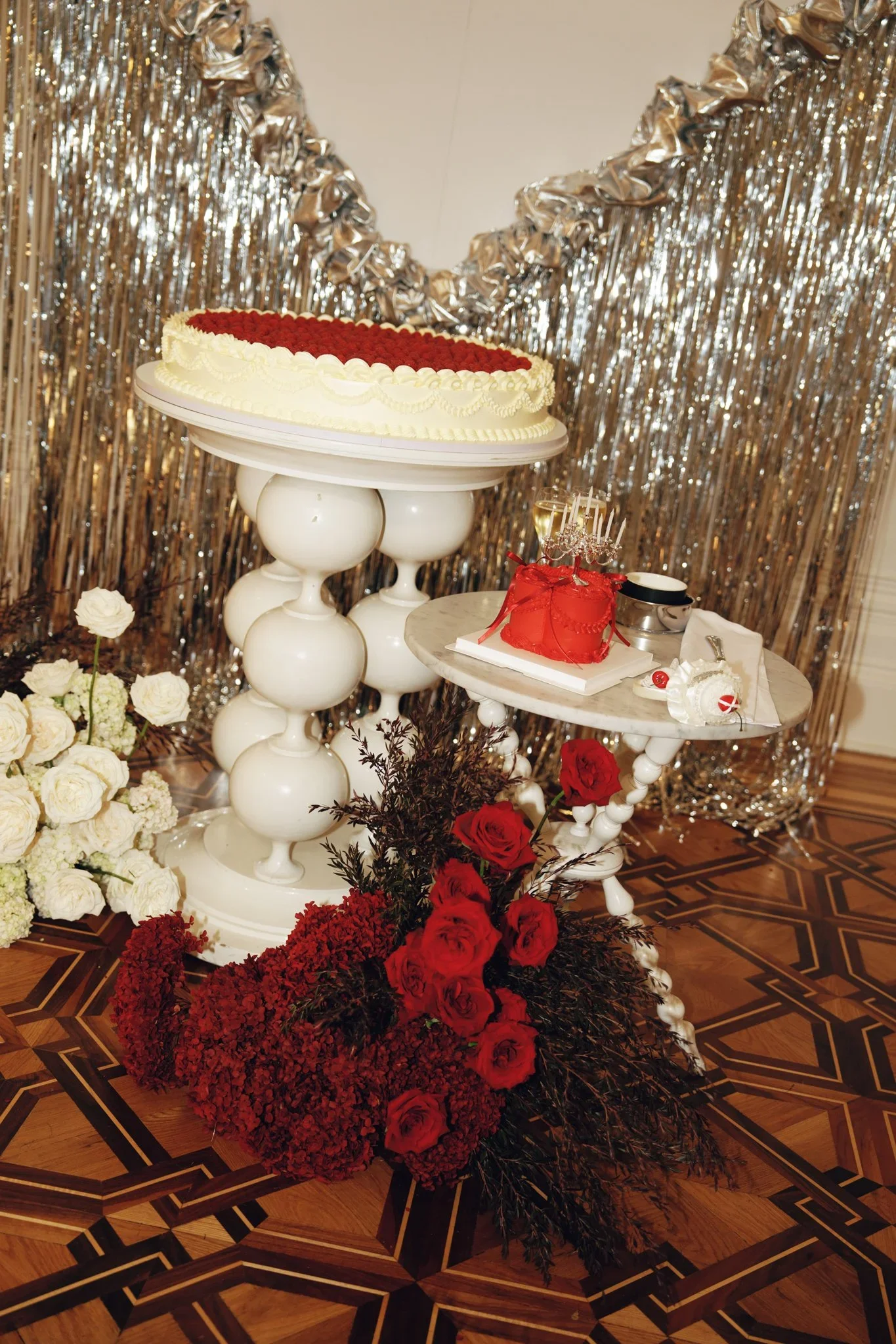 A decorated celebration setup with a large cake on a white pedestal, surrounded by red, white, and dark floral arrangements, with a silver foil curtain backdrop and smaller tables with gifts and flowers.