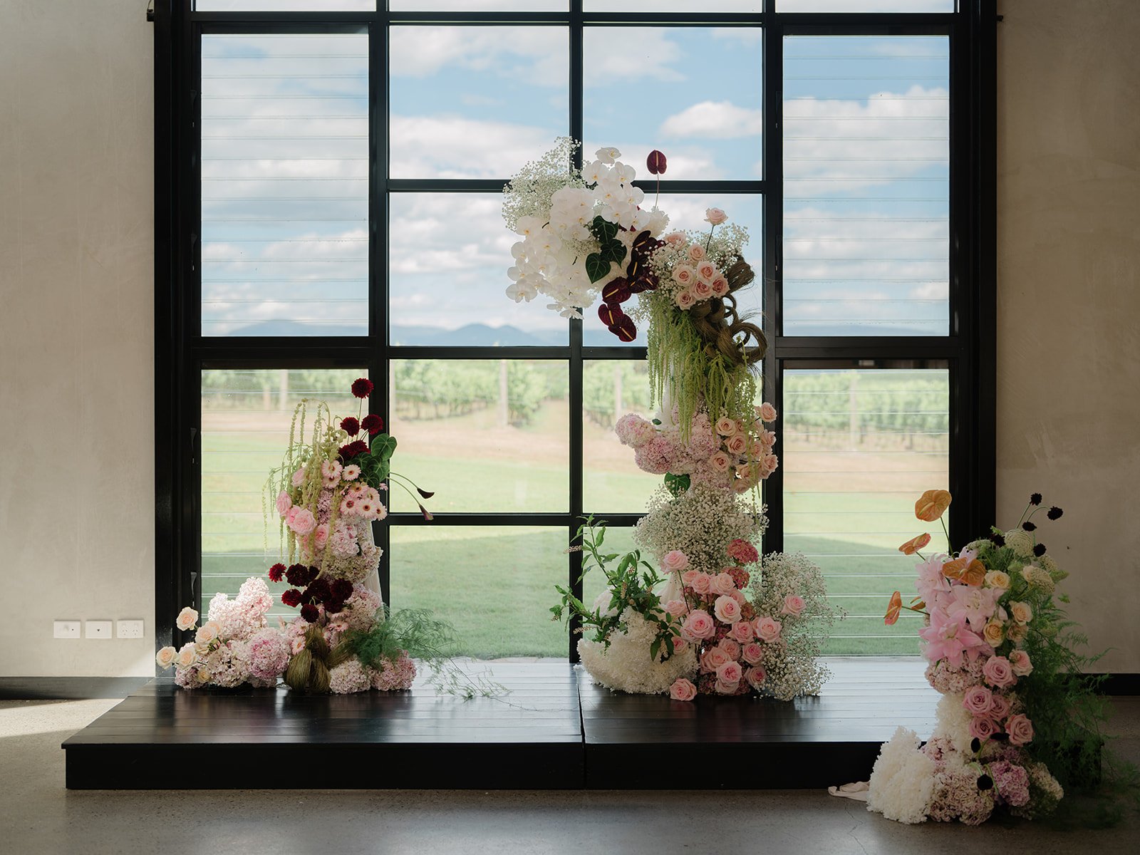 Arranged floral display with pink and white roses, orchids, and greenery in front of a large black framed window showing a green landscape and blue sky.