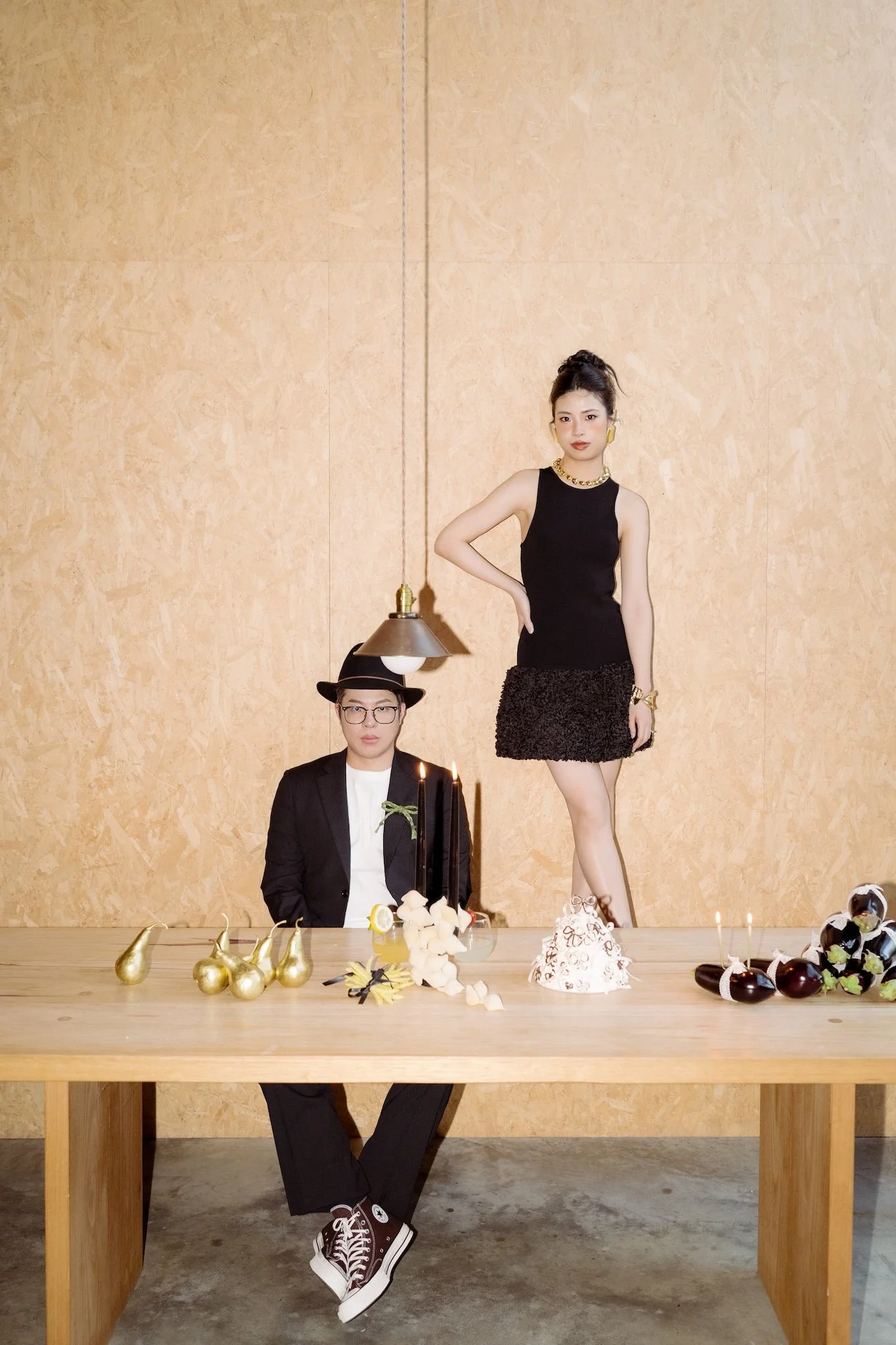 Two people at a celebration table, one man sitting with black sneakers and a black suit, a woman standing in a black dress with gold accessories, colorful cakes, and gold and black party decorations on the table.