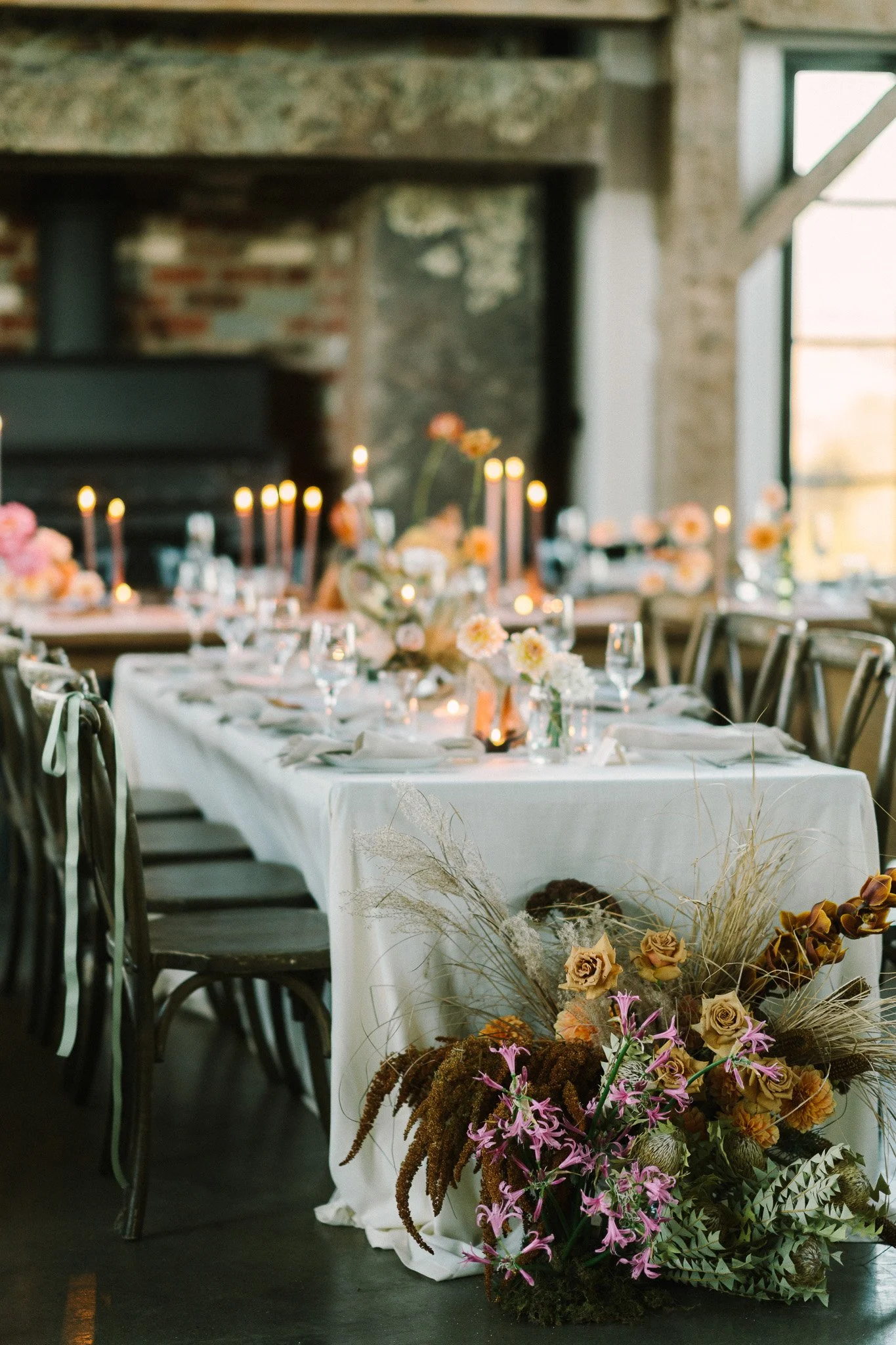 An indoor event space decorated with a long table set for a celebration, featuring floral centerpieces, candles, and elegant tableware, with a backdrop of brick and stone walls and large windows.