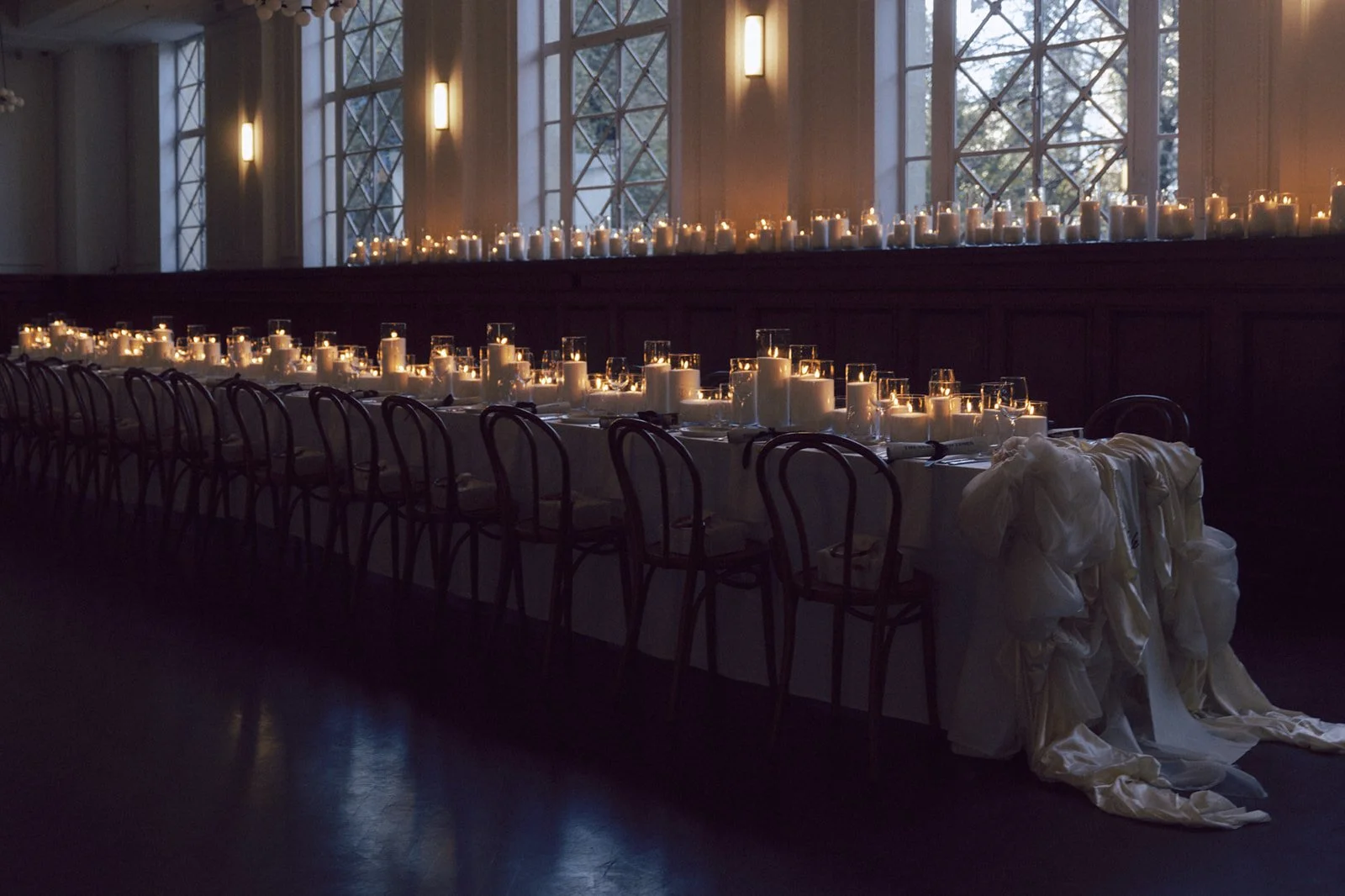 Long dining table set with numerous candles in glass holders and white cloth napkins, arranged in an elegant event space with large windows and soft indoor lighting.