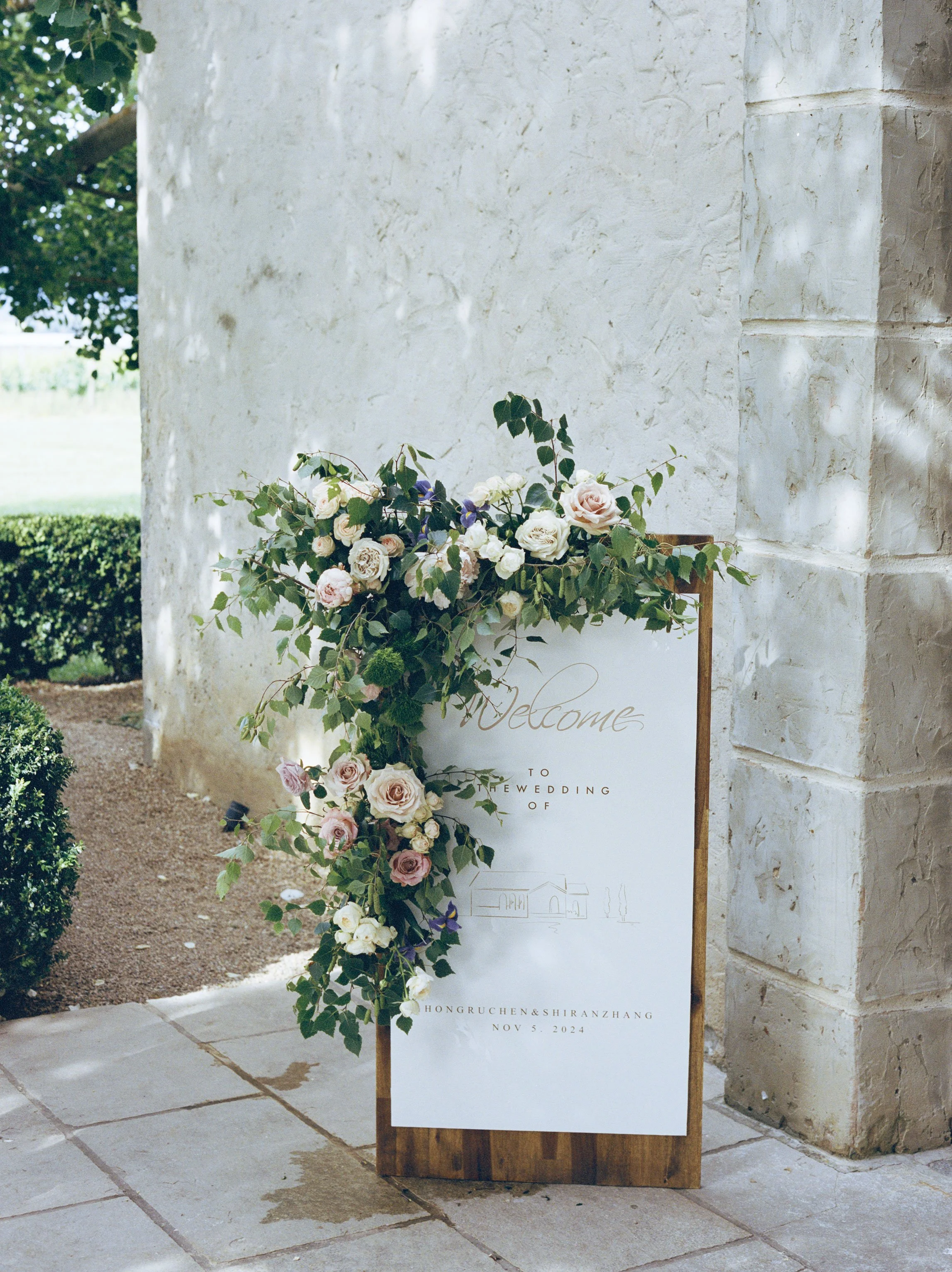 A white wedding welcome sign decorated with a floral arrangement of roses and greenery, standing on a wooden easel outside a stone building, with the date November 5, 2024, and the location Hongruchen & Shiran Zhang visible on the sign.